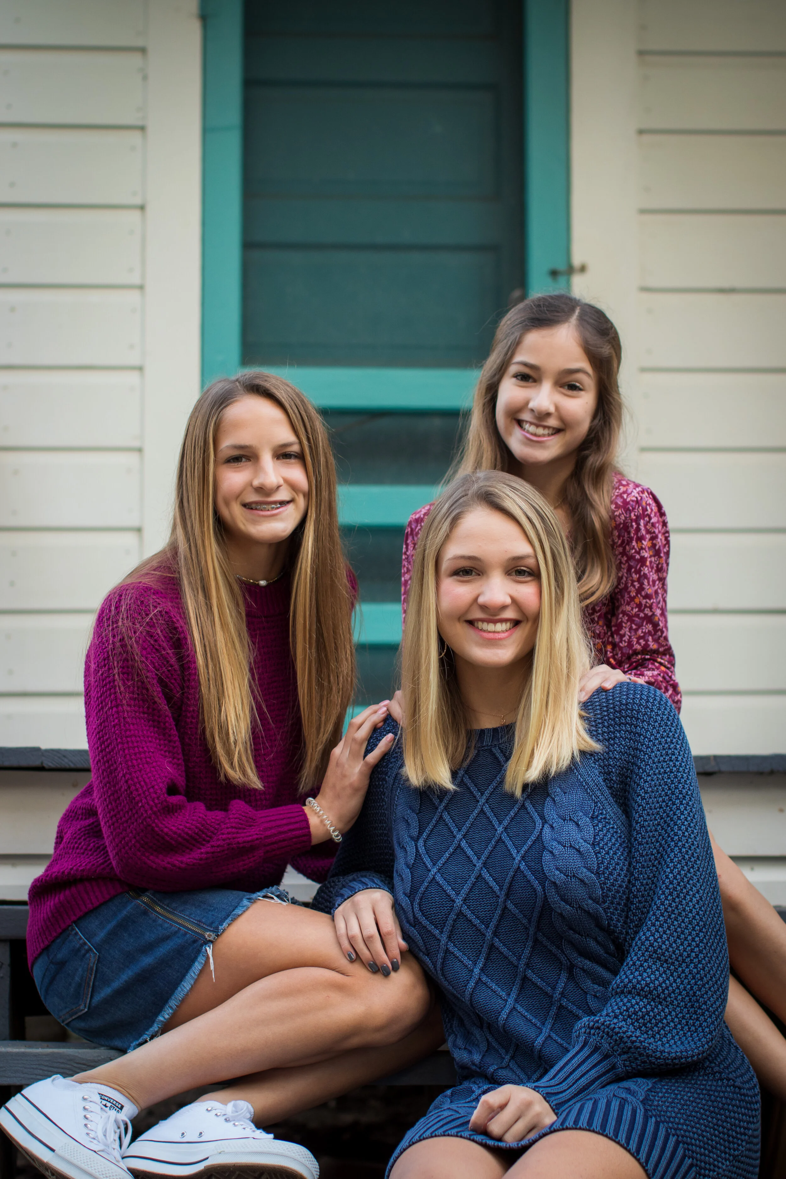 Four young women smiling and sitting together on the steps of a house with cream-colored siding and teal window trim.