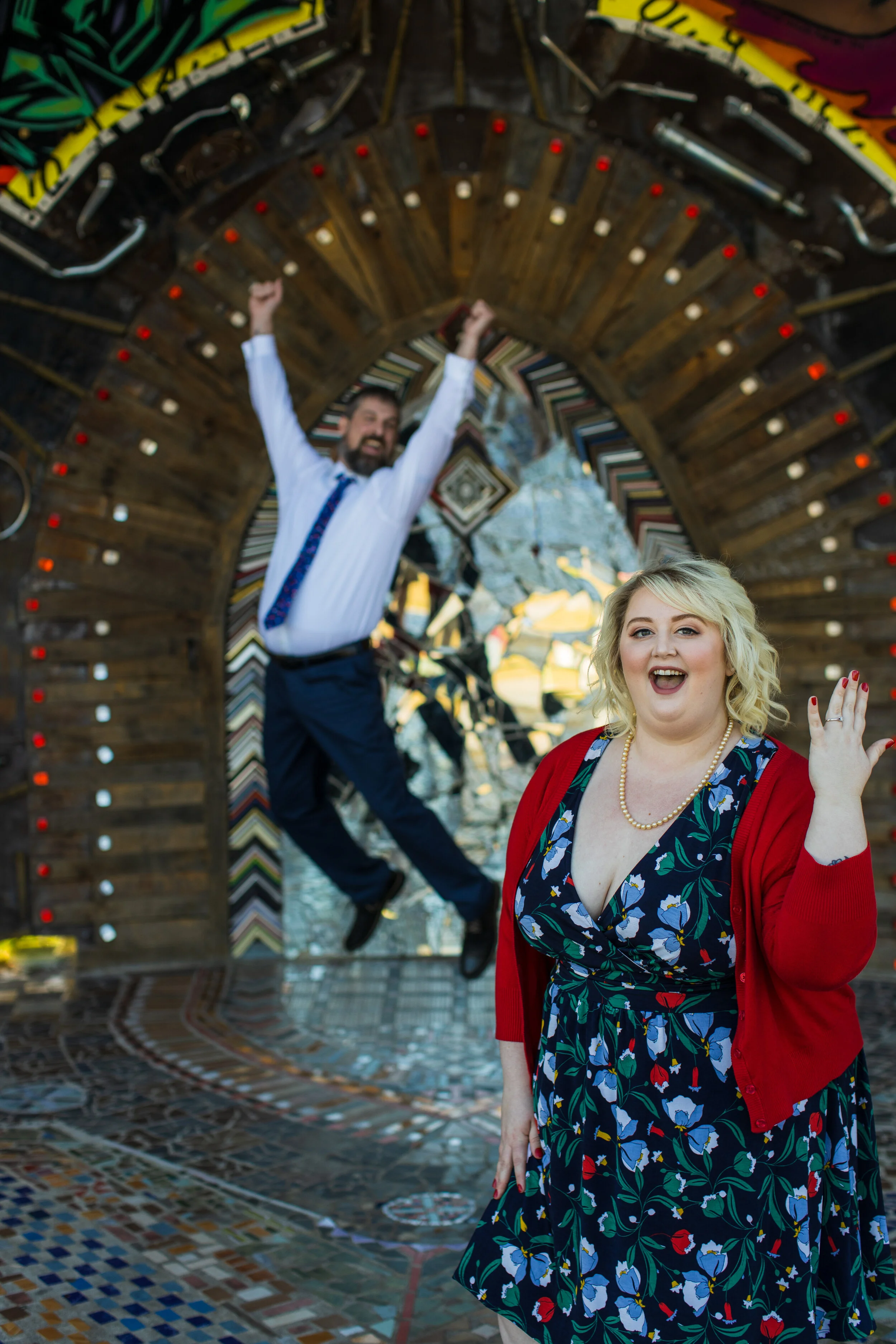 Two people, a man and a woman, are inside a tunnel made of various colorful book covers arranged in an arch. The man, dressed in a white shirt and blue tie, is jumping with his fists raised, and the woman, wearing a dress with a floral pattern and a 