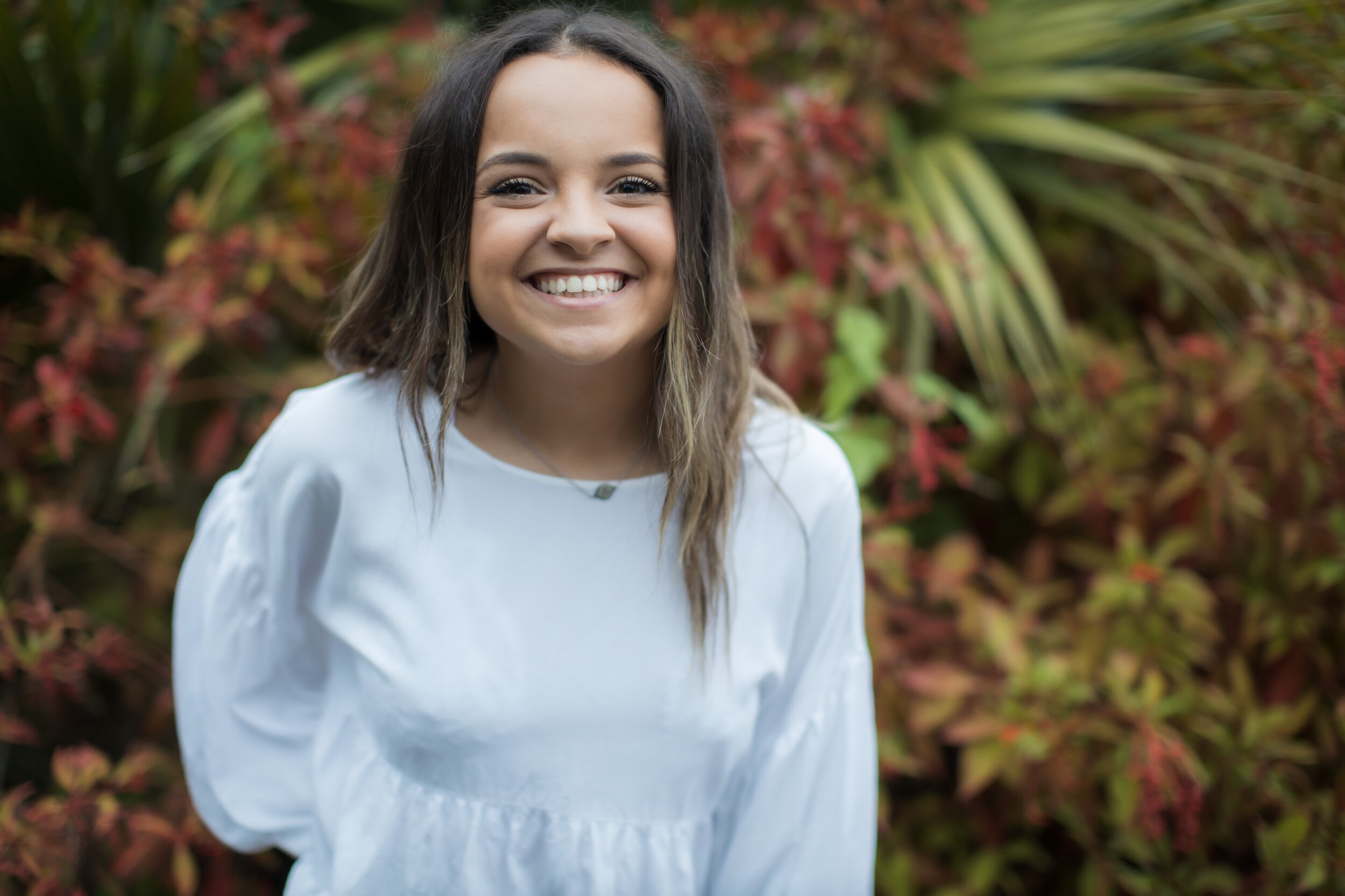 A young woman smiling outdoors in front of colorful foliage.