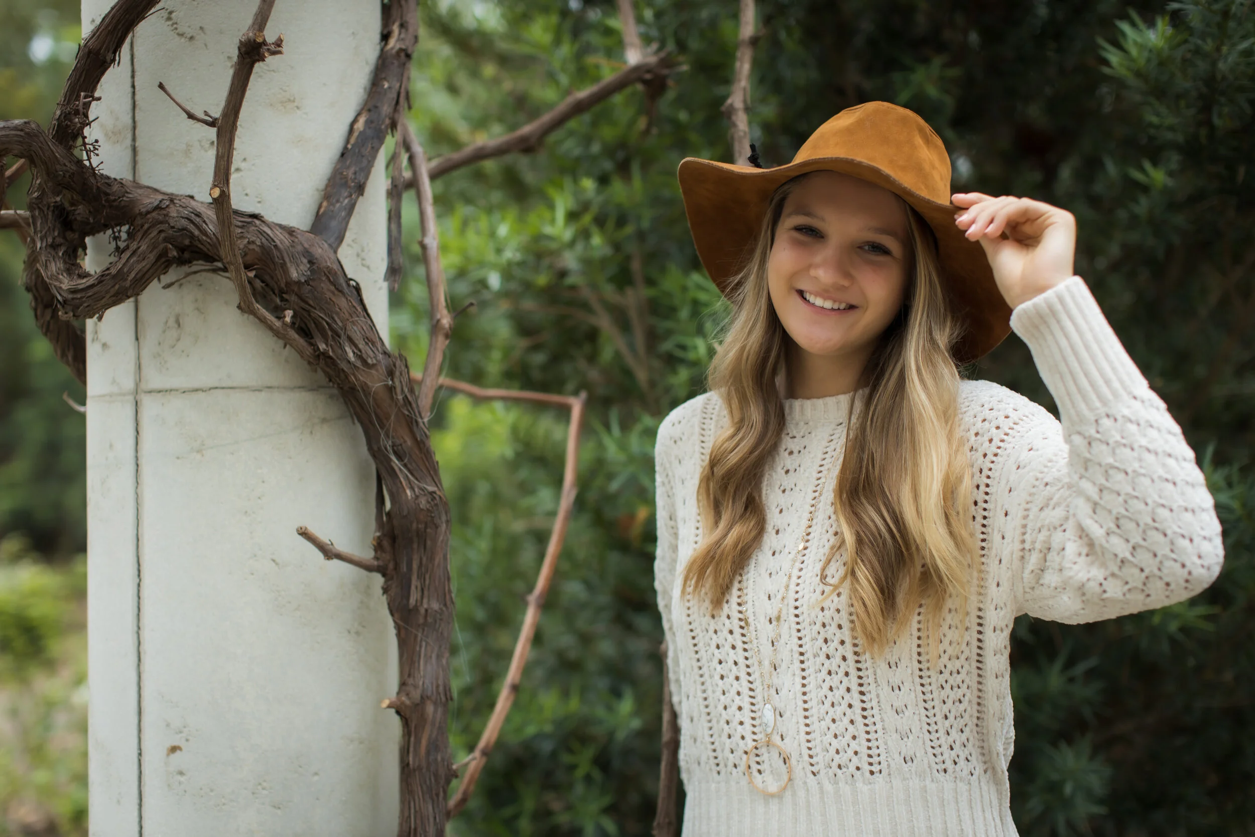 A young girl with long blonde hair wearing a beige sweater and a brown hat, smiling while standing outdoors next to a white post with vine climbing on it, surrounded by greenery.