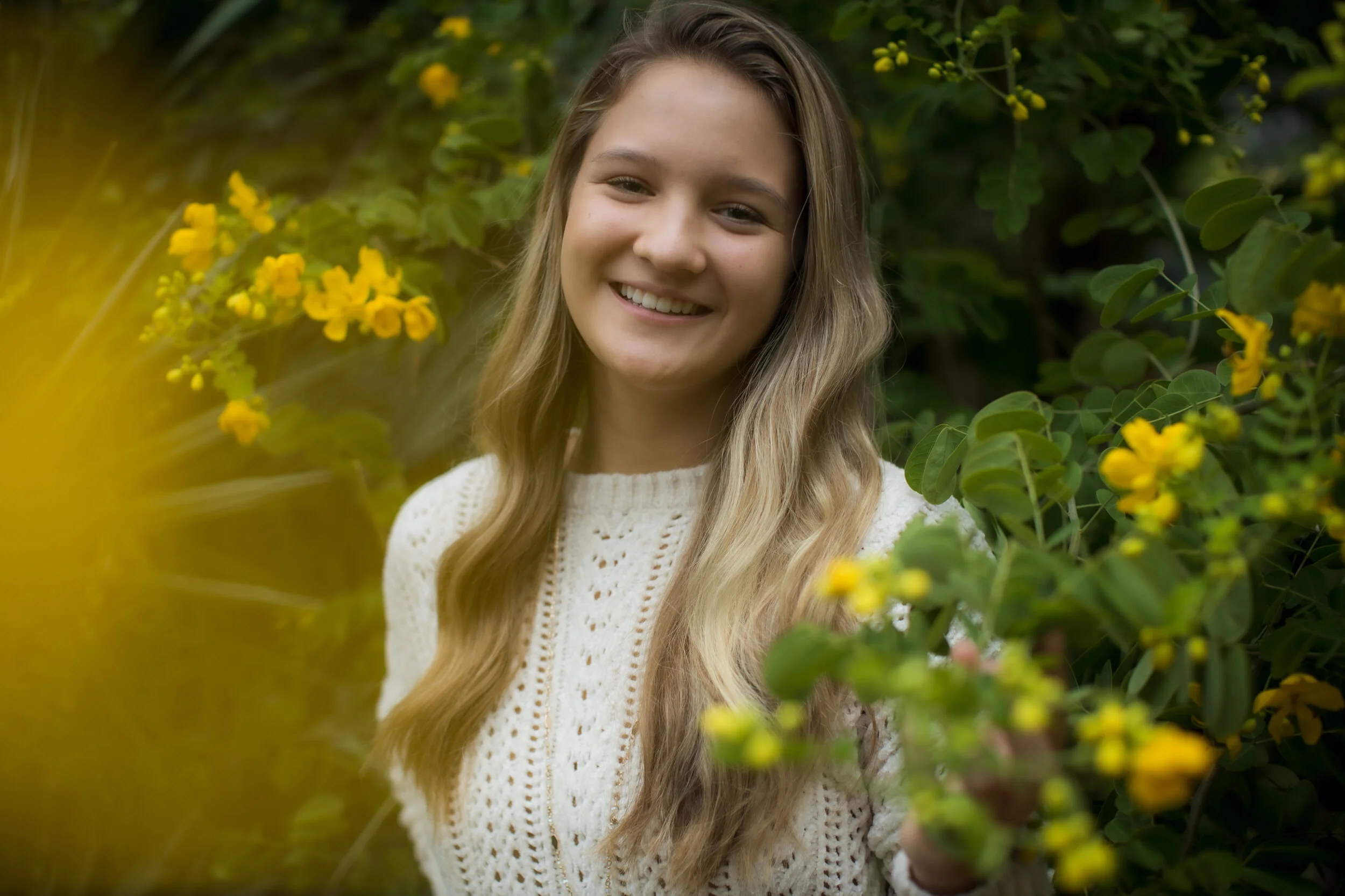 A young woman with long blond hair smiling among green foliage and yellow flowers.