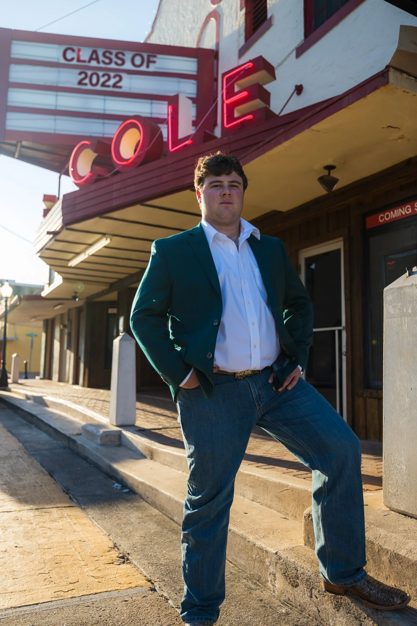 A young man in a teal blazer, white shirt, and jeans stands confidently outside a restaurant with a neon "COLE" sign, with a sign in the background announcing the 2022 graduating class.