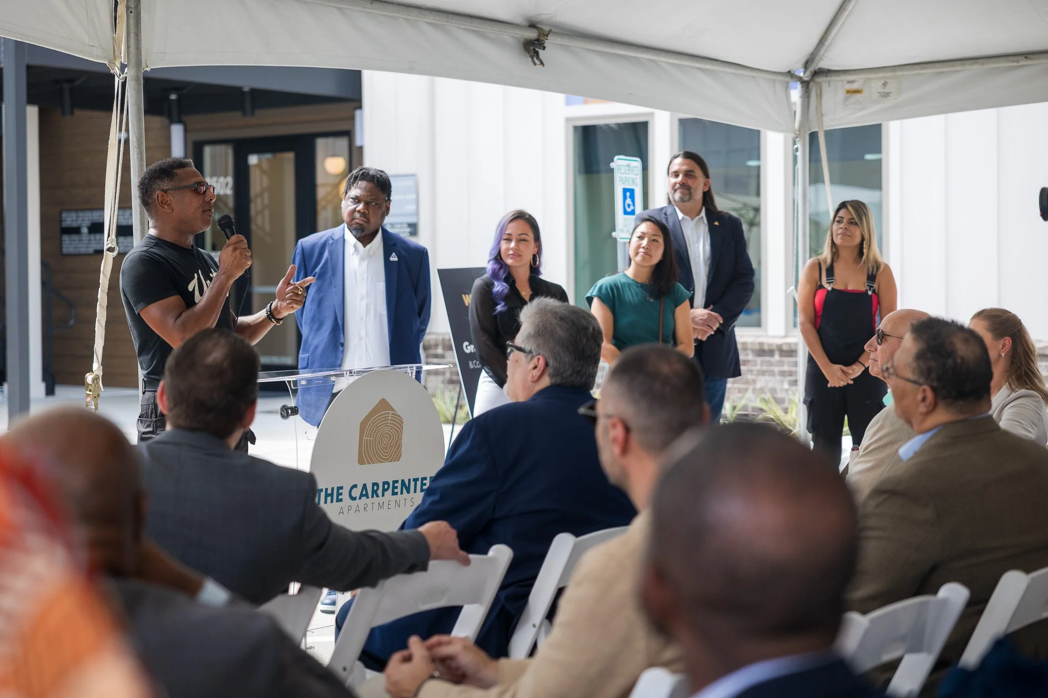 A group of people gathered under a tent at an outdoor event. One man is speaking into a microphone, while others stand nearby listening. The audience is seated and paying attention.