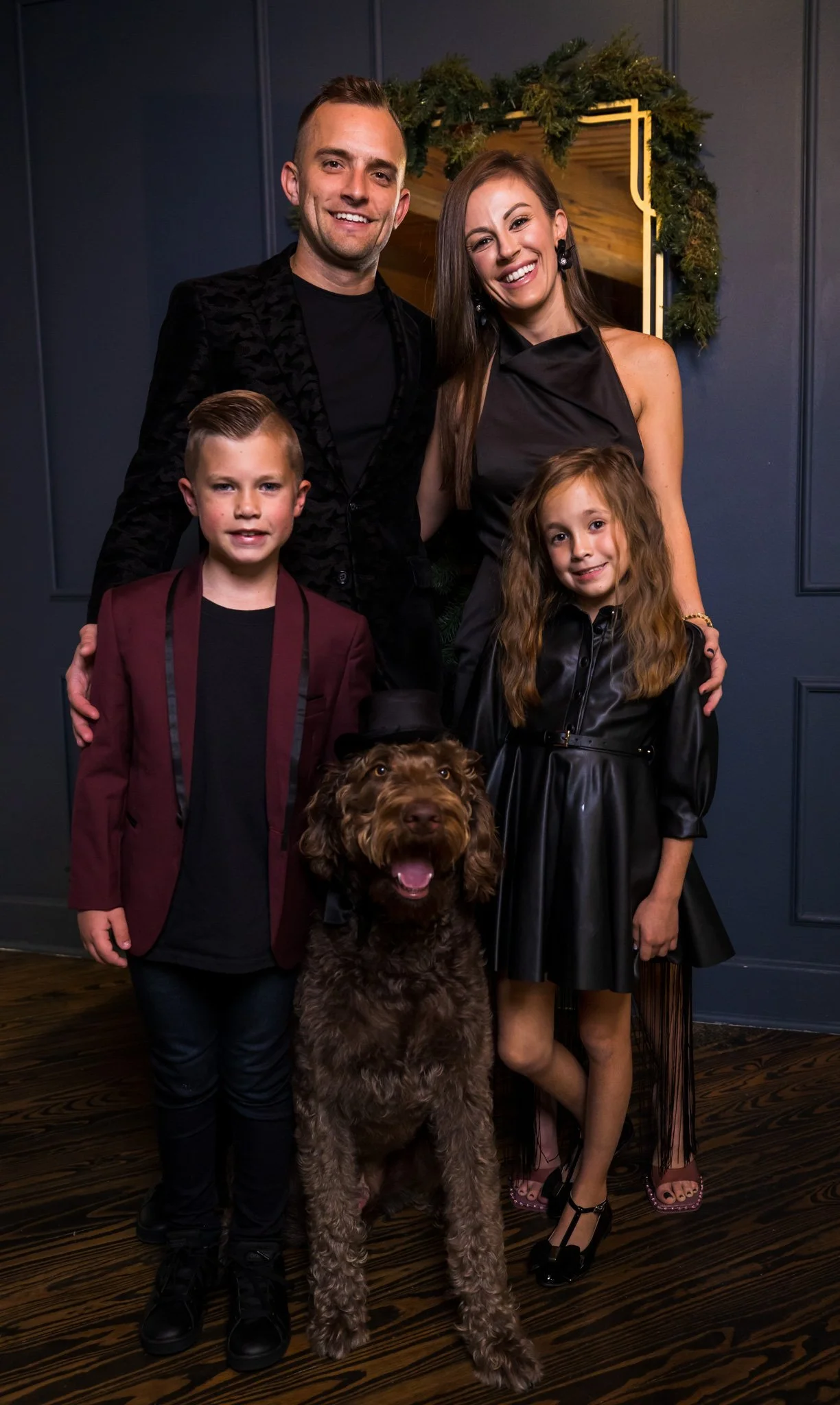 A group of five people and a dog posing for a photo. There are two men, two women, a large brown dog, and a young boy with blond hair. They are indoors with dark blue walls and a mirror decorated with Christmas greenery in the background.
