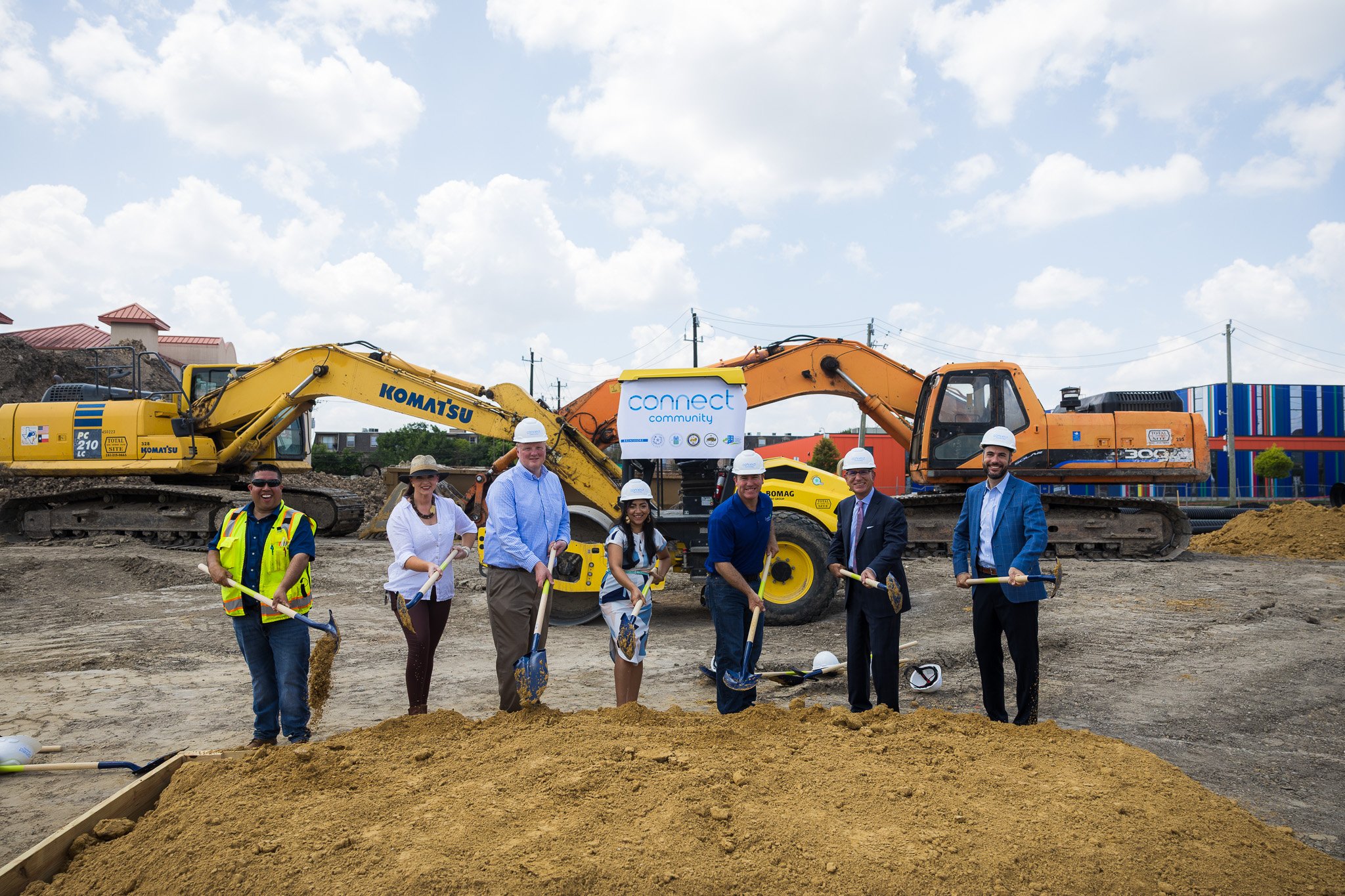 Group of people at a construction site holding shovels, with construction equipment and machinery in the background.