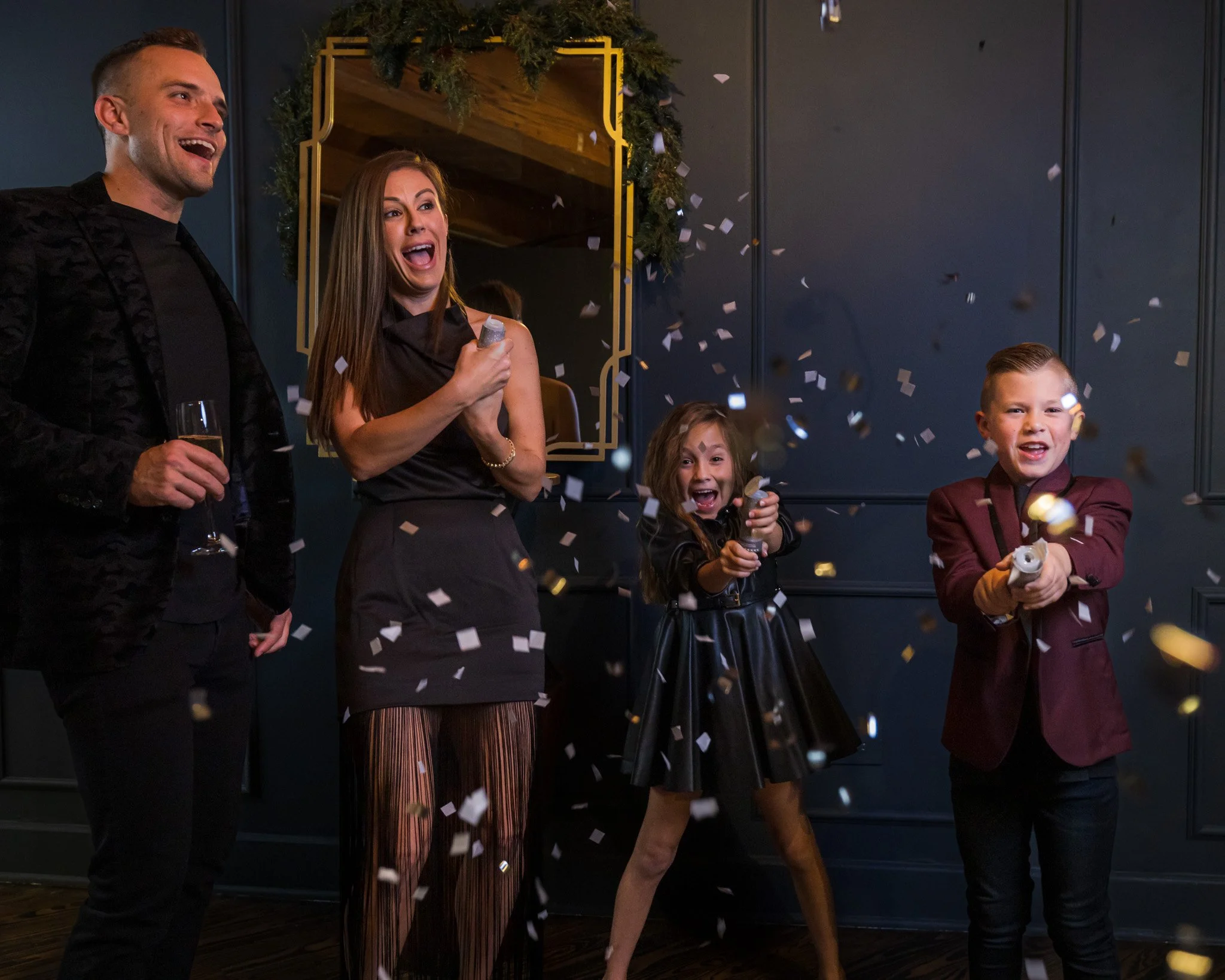 Group of people celebrating with confetti, including a man with a champagne glass, a woman holding a confetti popper, and children in festive attire, indoors with dark walls and holiday decor.
