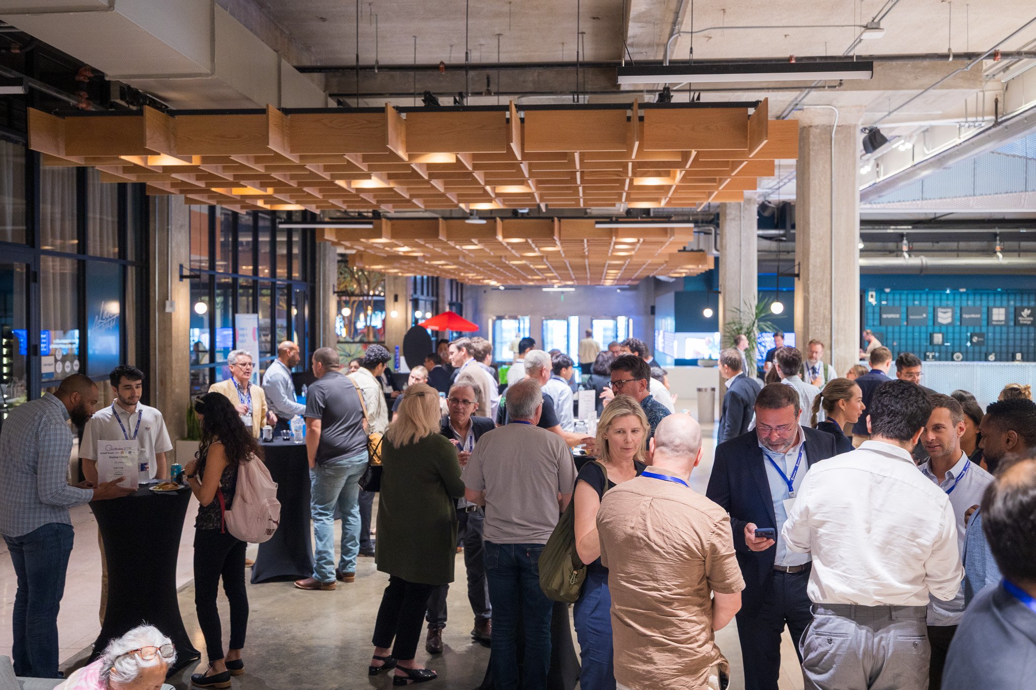 A crowded indoor event with many people gathered around tables, talking and socializing under a wood-panel ceiling with industrial elements.