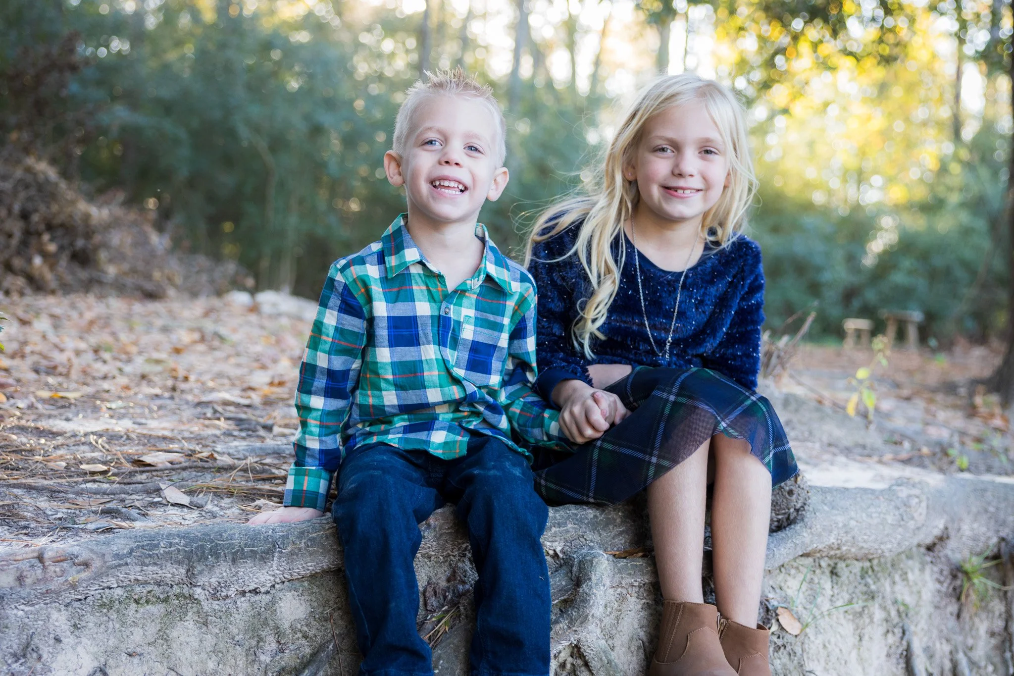 Two children, a boy and a girl, smiling and sitting on a log in a wooded area with sunlight filtering through trees.