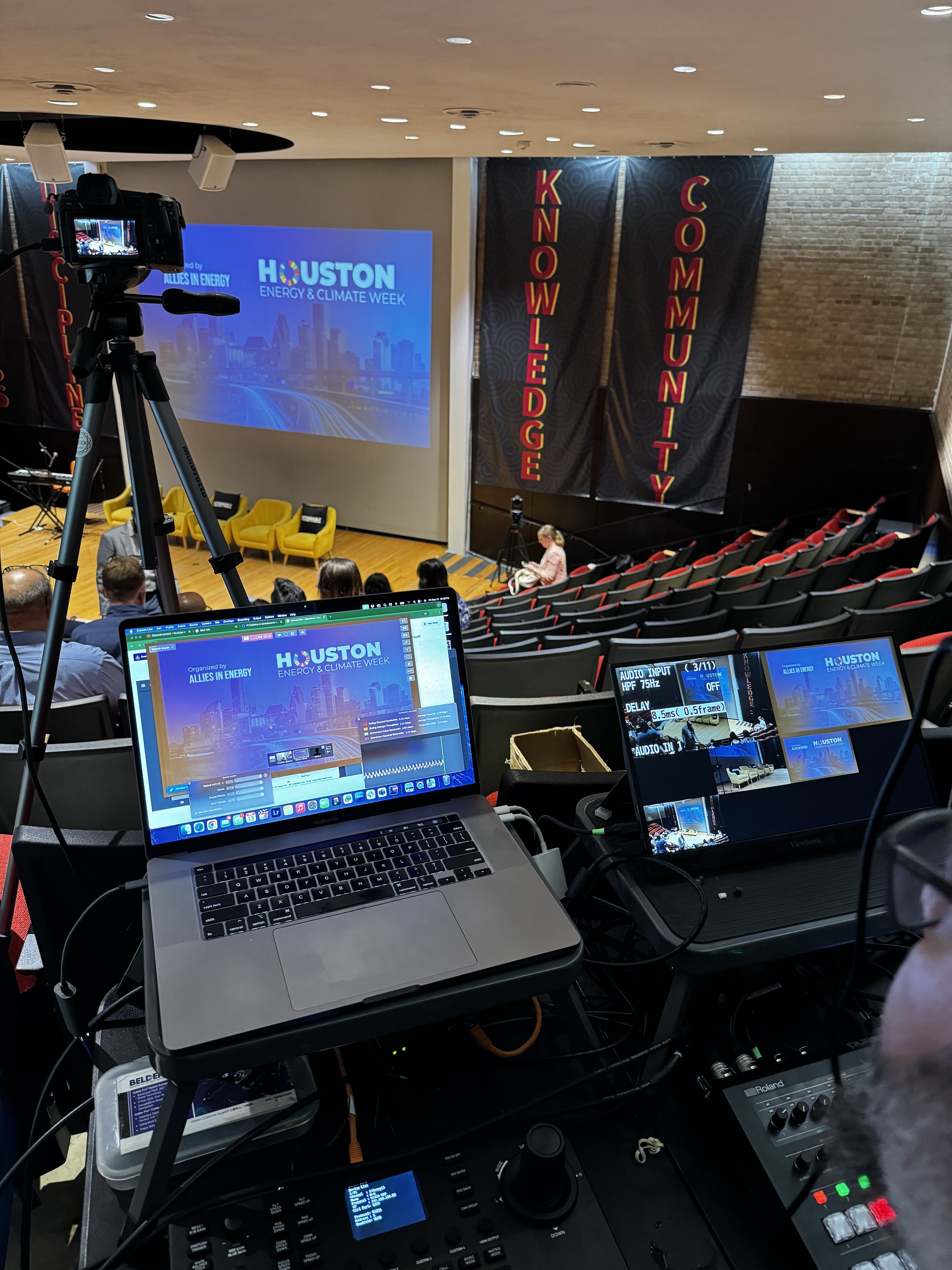 A conference room with a large screen displaying the Houston Energy & Climate Week logo and text, yellow chairs on stage, and a camera capturing the event. In the foreground, there are laptops, microphones, and audio equipment on tables, and banners reading 'Knowledge', 'Community', and 'Showledge'.