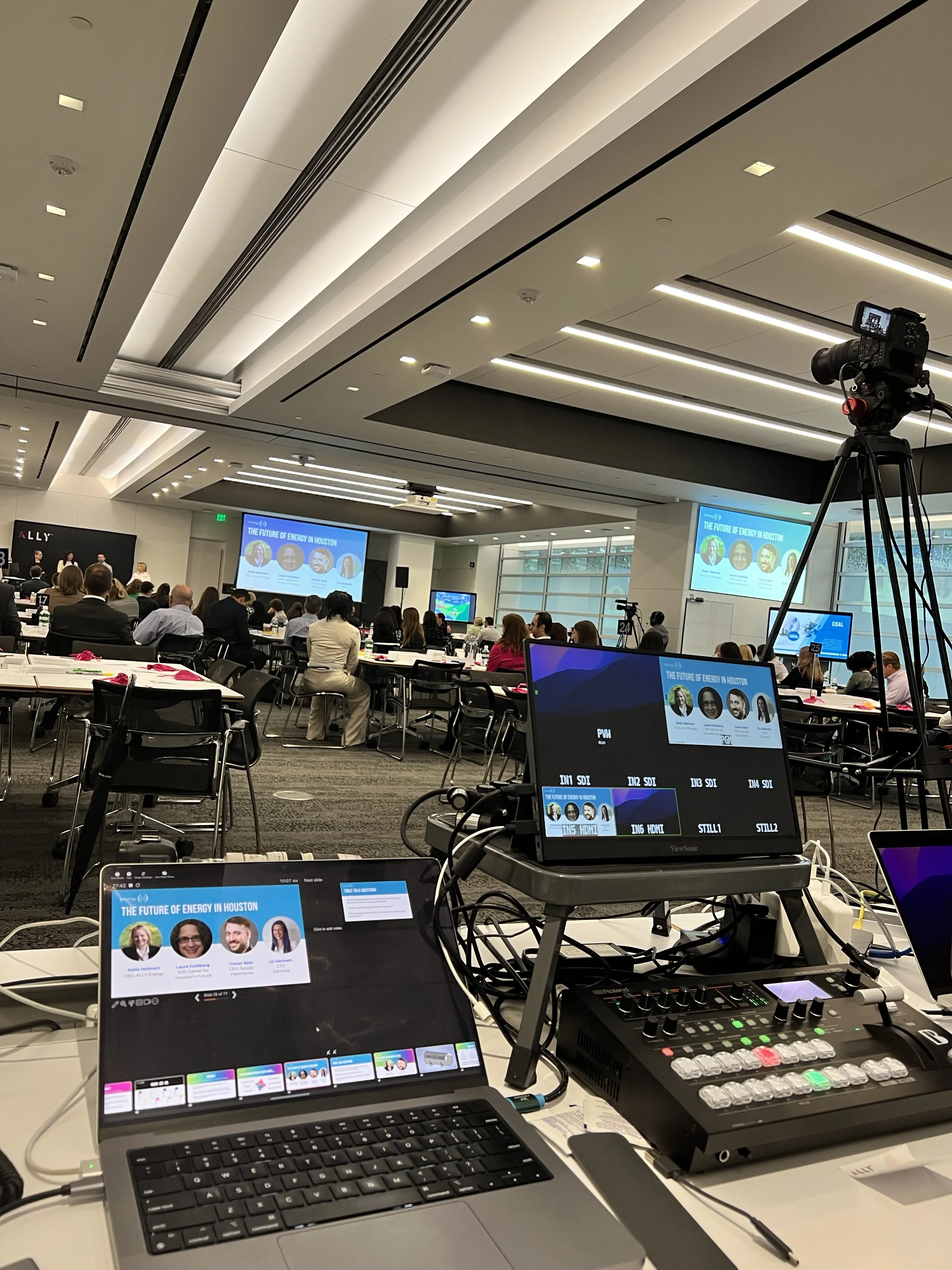 Conference room with multiple people seated at tables, large screens displaying a presentation titled "The Future of Energy in Houston," and various electronic equipment including laptops, monitors, and a camera on a tripod.