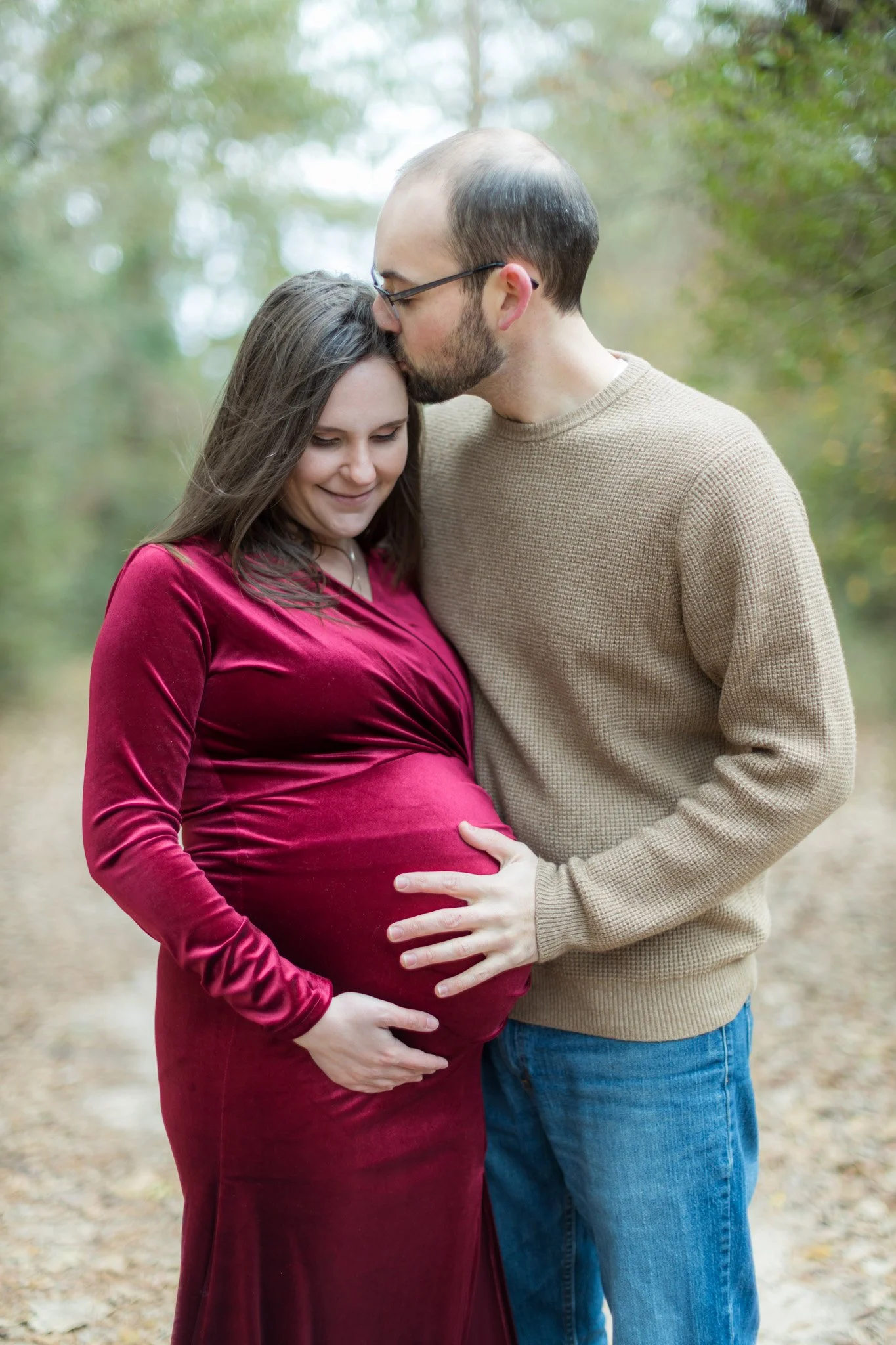 A pregnant woman in a red velvet dress with long brown hair smiling and a man in a tan sweater with glasses kissing her forehead, standing outdoors with blurred green and yellow trees in the background.