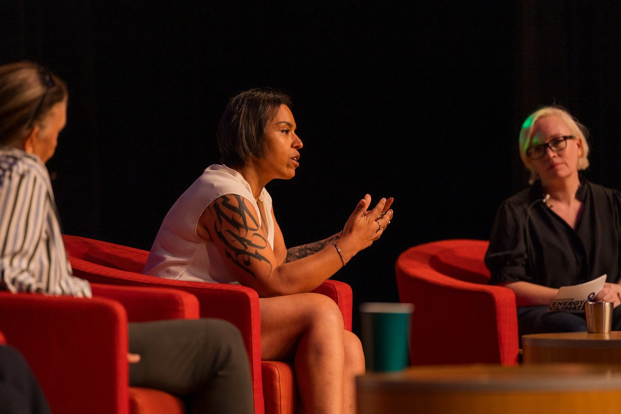 Three women engaged in a discussion seated on red chairs against a dark background, with a woman in the center speaking.