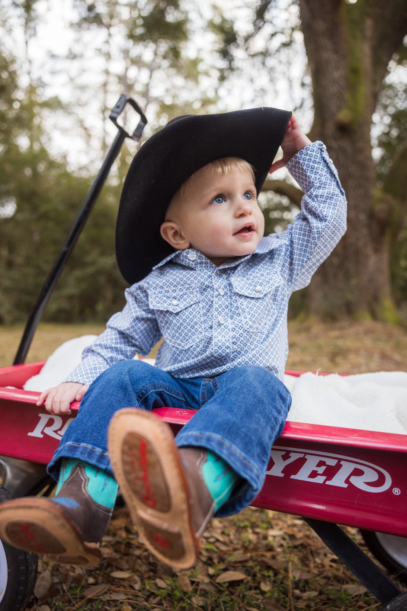 Young boy sitting in a red wagon with a black hat, outdoors in a wooded area, wearing a patterned shirt and jeans, looking up while holding his hat.