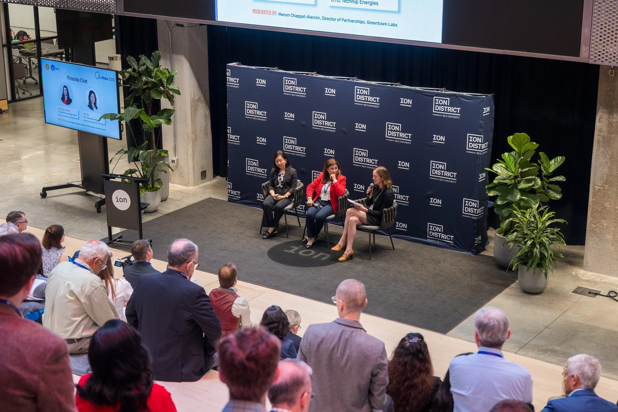Three women seated on a stage participating in a panel discussion at the Ion District event. Audience members are seated in front of them, and there are two large potted plants on either side of the stage. A large screen behind the panel displays the Ion District logo repeatedly, with the event branding. A smaller screen on the left shows a digital fireside chat with two speakers' photos and names.