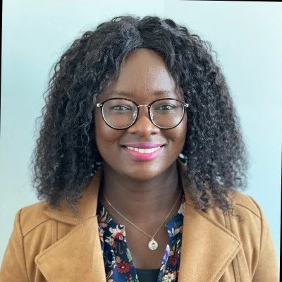Portrait of a woman with curly black hair, wearing glasses, a tan jacket, and a patterned blouse, smiling against a light blue background.