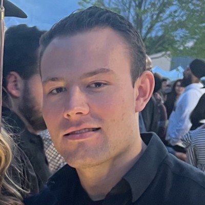 A young man with short, dark hair and light skin, smiling and wearing a black shirt, outdoors during daytime with trees and a crowd in the background.