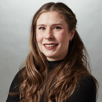 A young woman with long, wavy brown hair and light skin, wearing a black top, smiling in front of a light gray background.