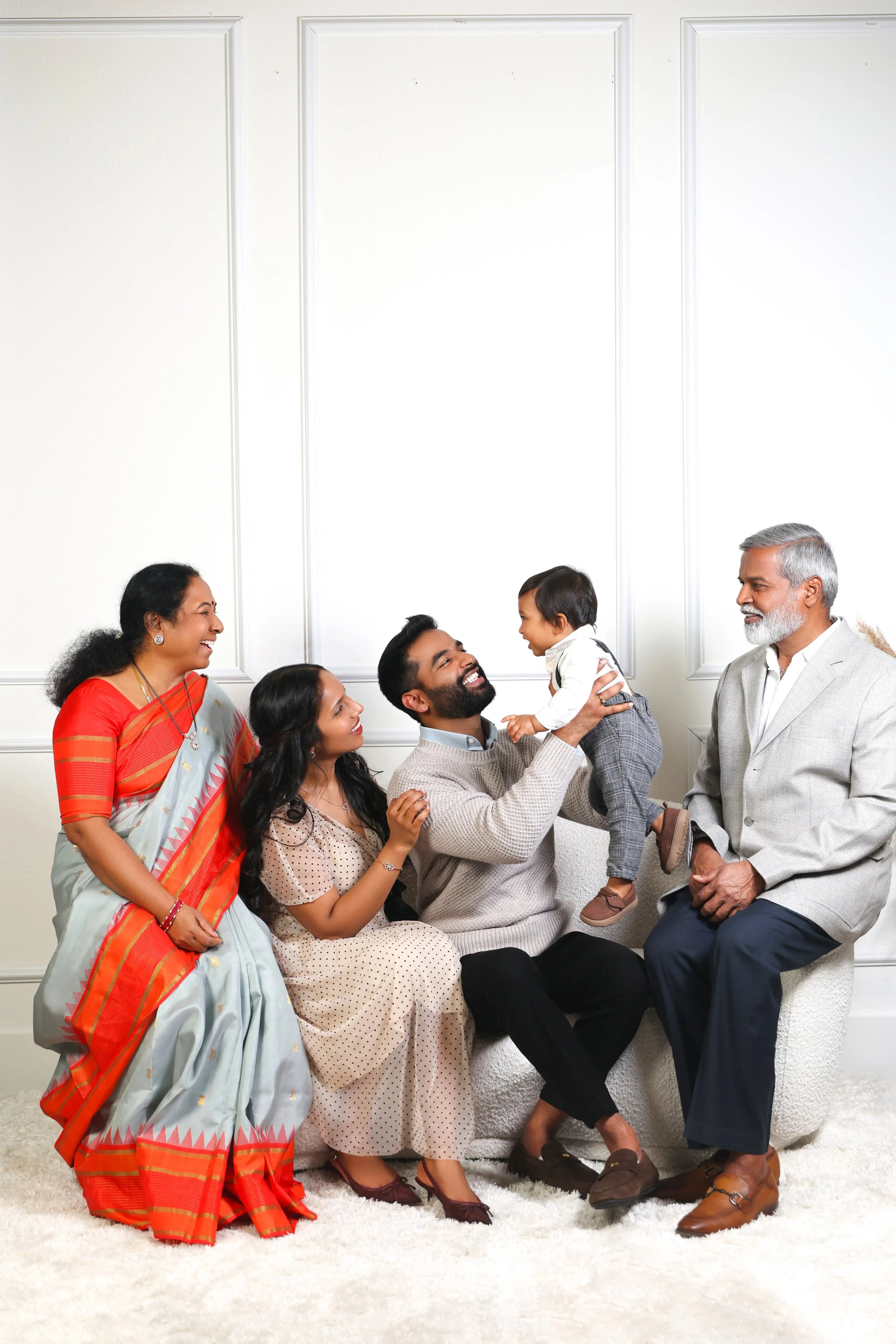 Family sitting on a white carpet in front of a white wall, with a woman in a red saree on the far left, a woman in a beige dress next to her, a man holding a young boy in his lap in the middle, and an older man in a gray suit on the right, all smilin