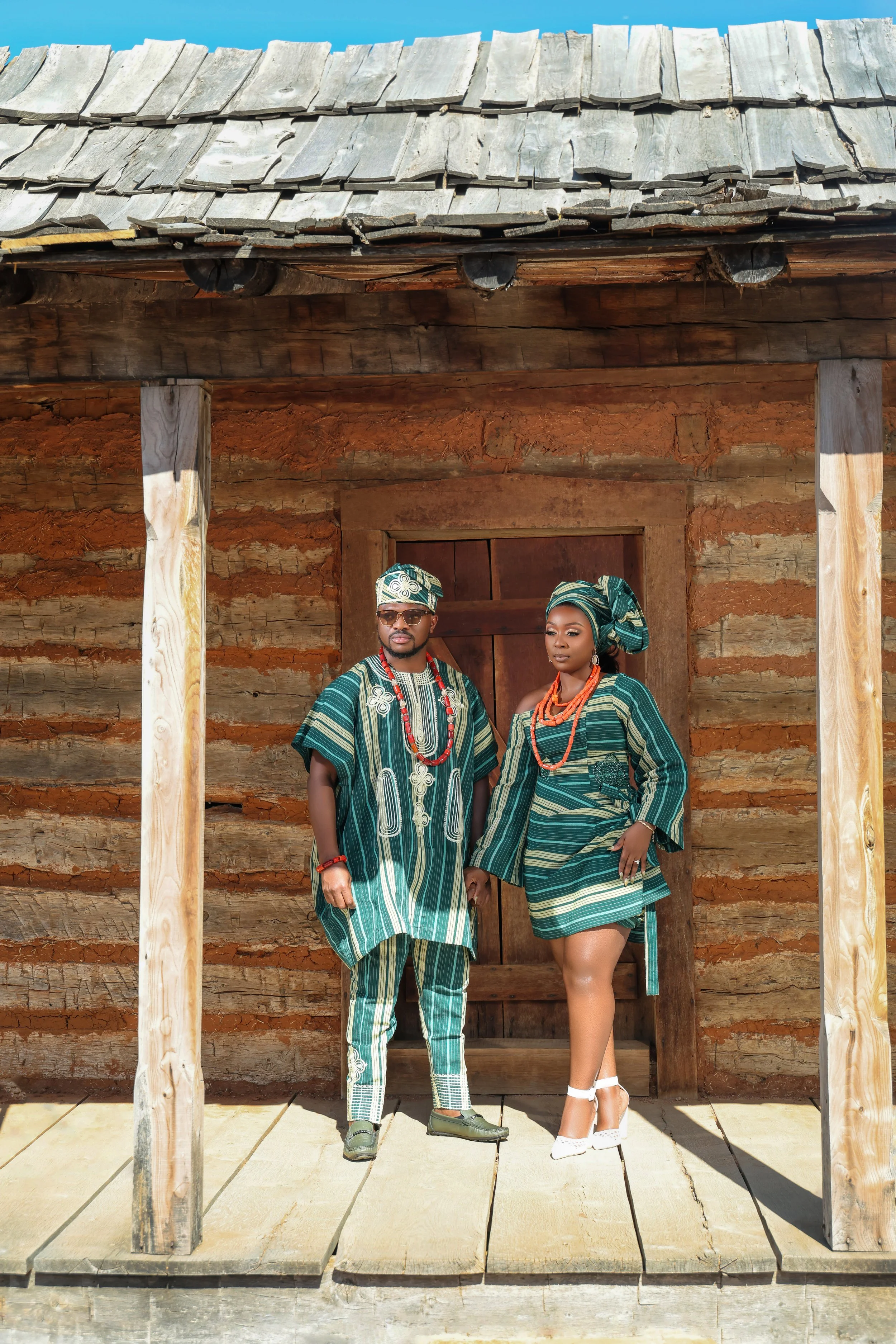 A man and woman dressed in traditional patterned clothing and head wraps, holding hands outdoors in front of a rustic wooden house.