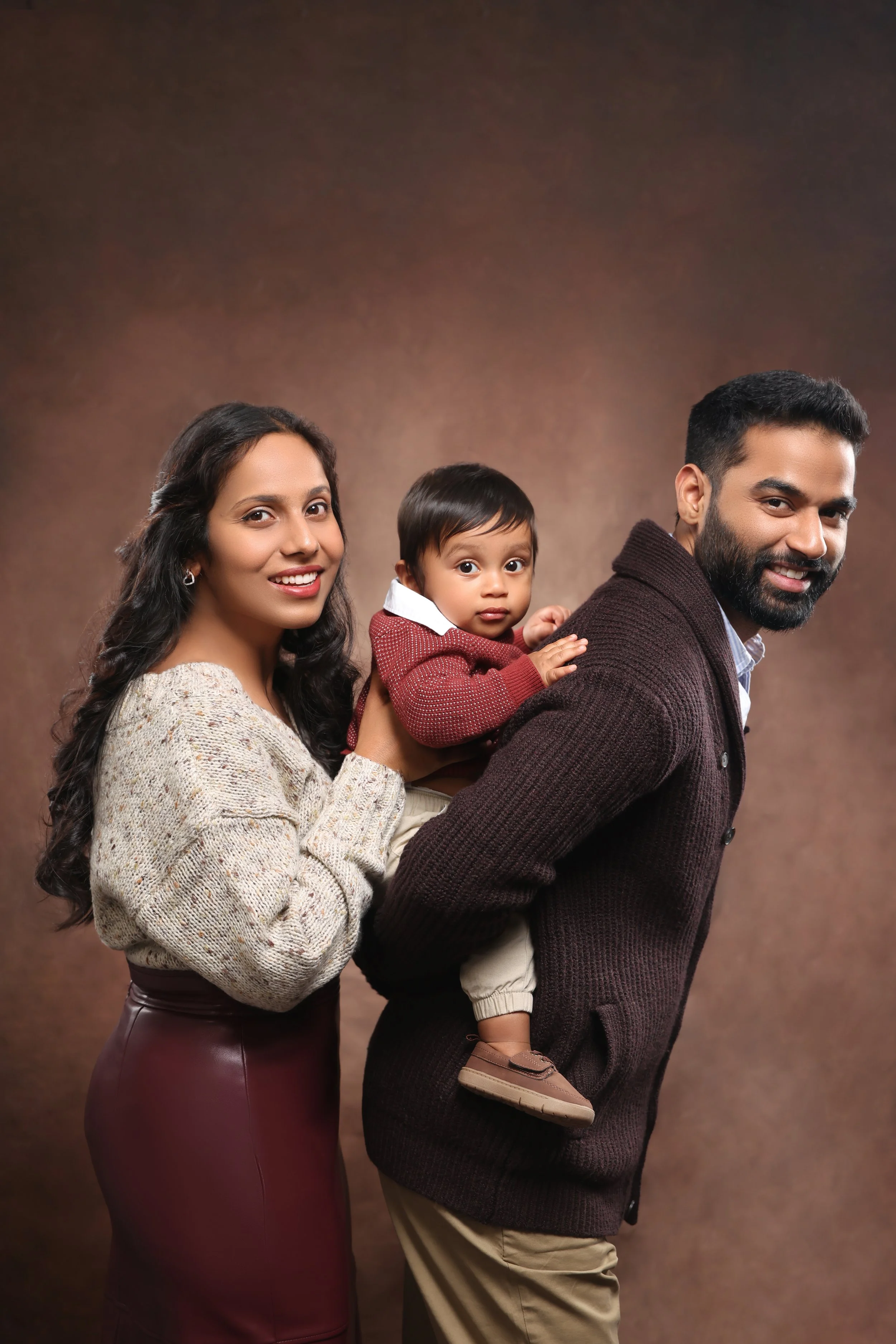 A family of three, including a woman with long dark hair in a sweater and skirt, a man with a beard in a cardigan, and a young child in a red sweater, posing together in a studio against a brown background.