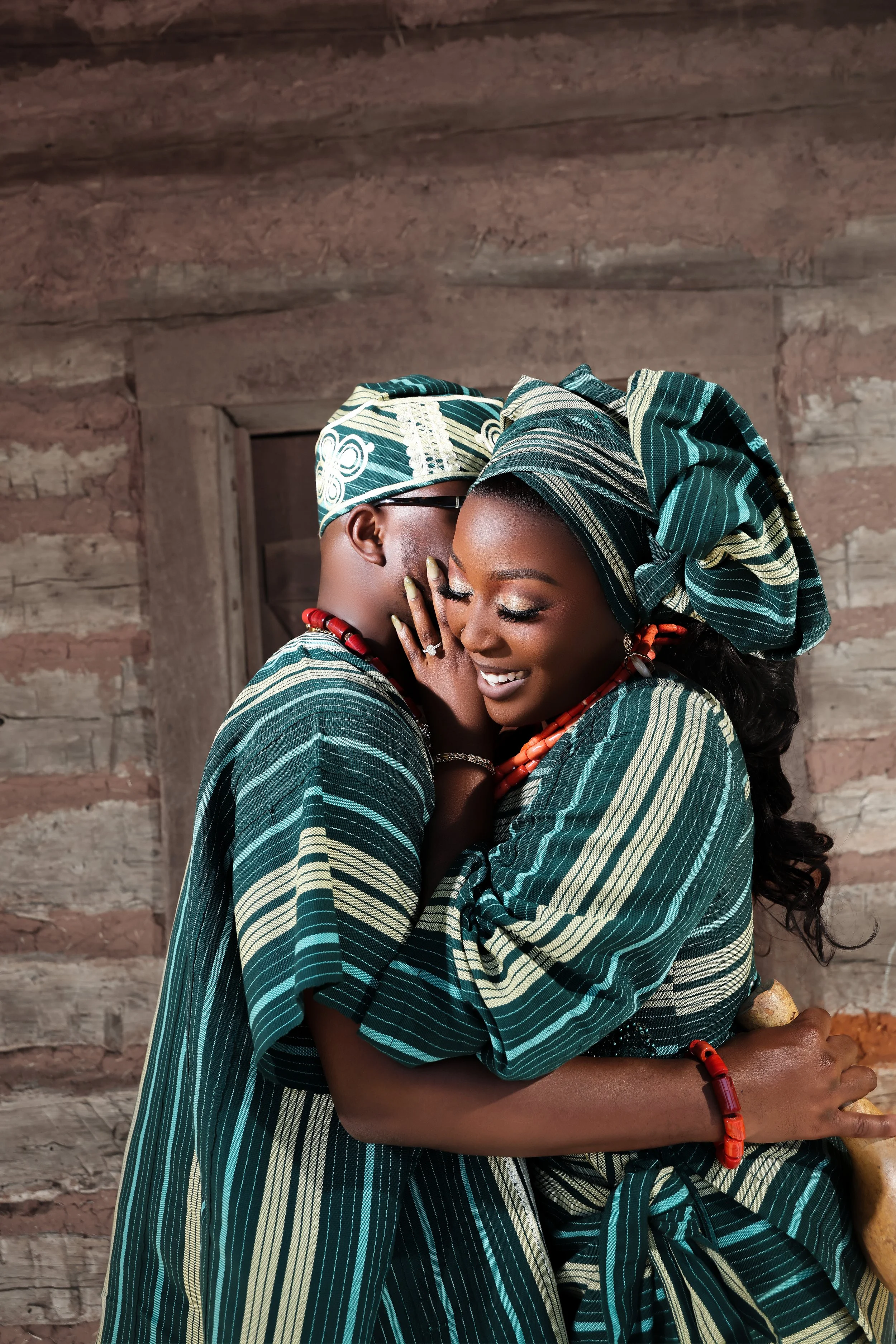 A smiling Black couple dressed in traditional African attire, embracing and sharing a joyful moment inside a rustic wooden building.
