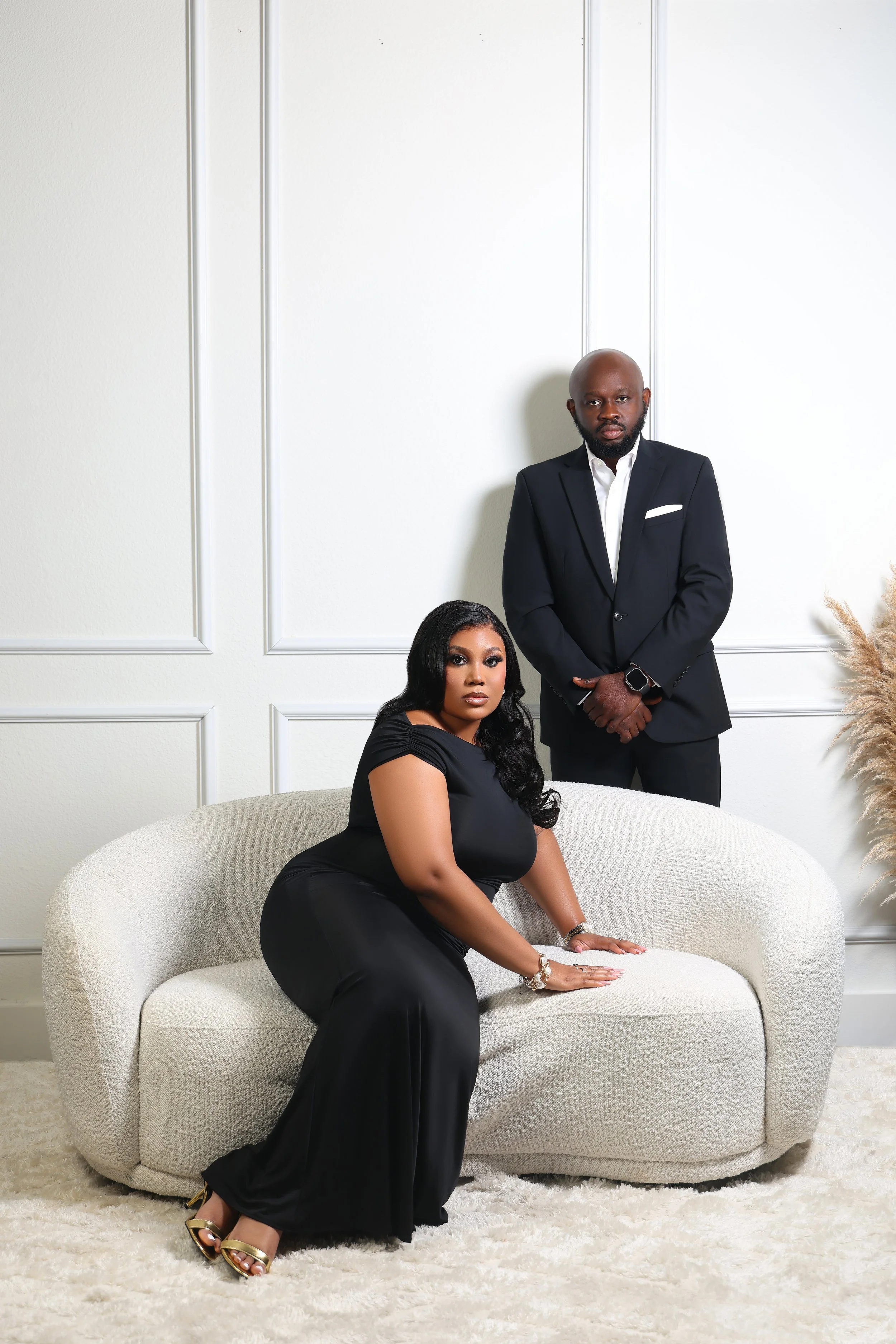 A woman in a black dress seated on a cream-colored textured sofa with a man standing behind her in a black suit against a white wall with decorative wall panels.