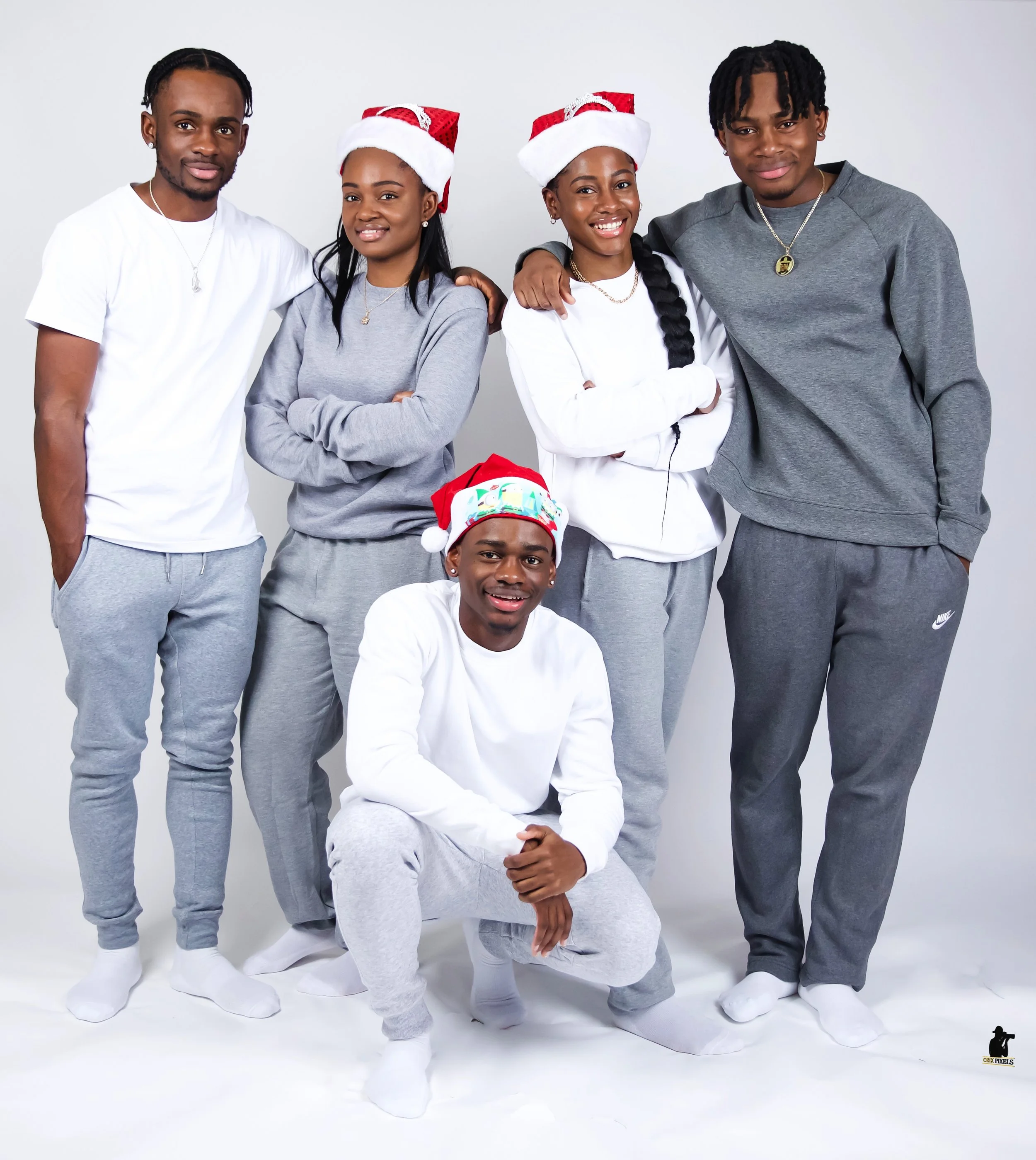 A group of six young people dressed in casual gray and white clothing, some wearing Christmas hats, posing together against a plain white background. One person is kneeling in front, four are standing, and two are wearing Santa hats.
