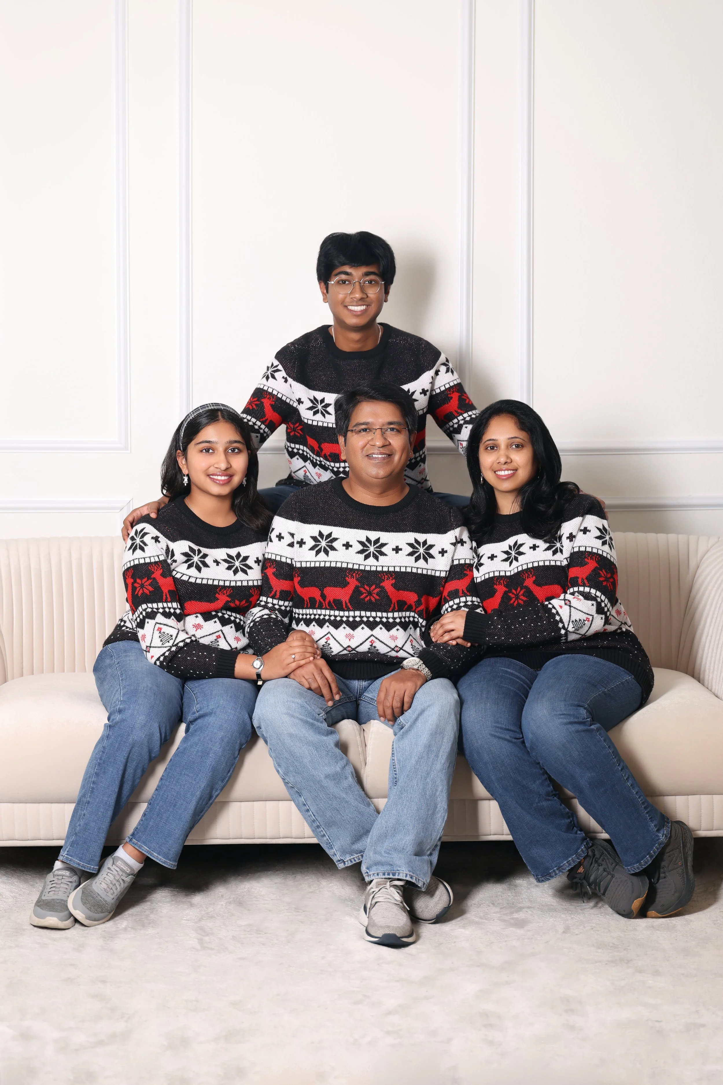 A family of five sitting and standing on a cream-colored sofa in a room with white wall paneling, all wearing matching Christmas sweaters with holiday patterns including reindeer and snowflakes.