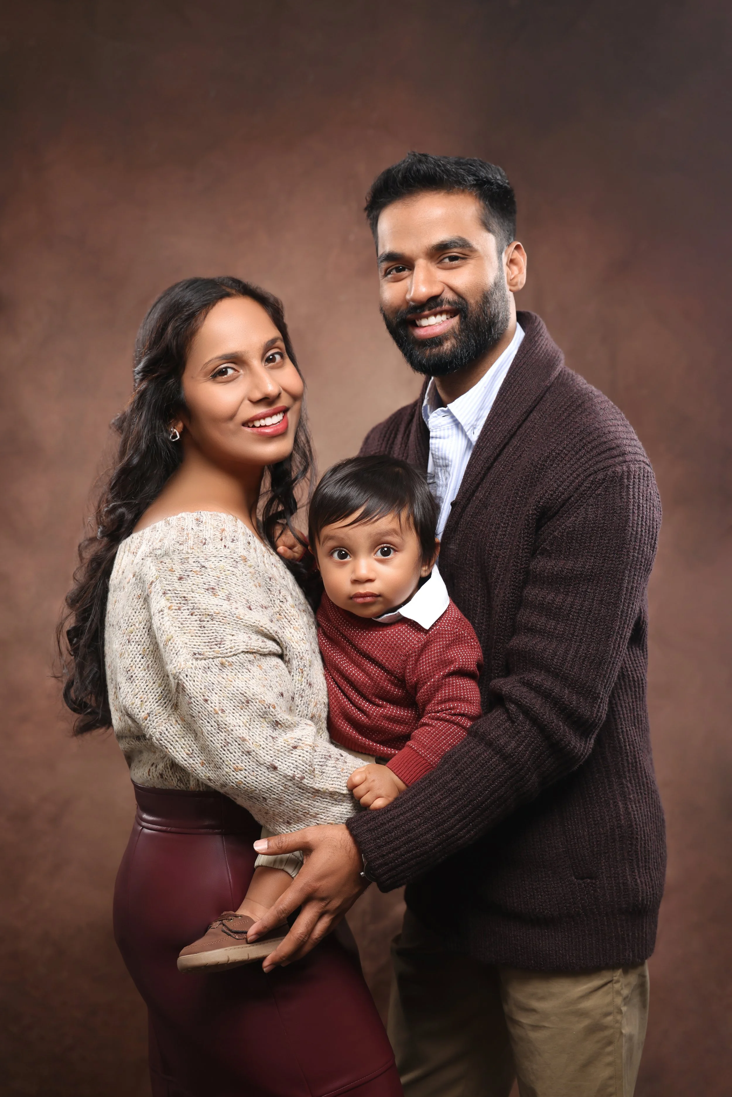 A happy family of three, including a woman, a man, and a young boy, posing together against a brown background.
