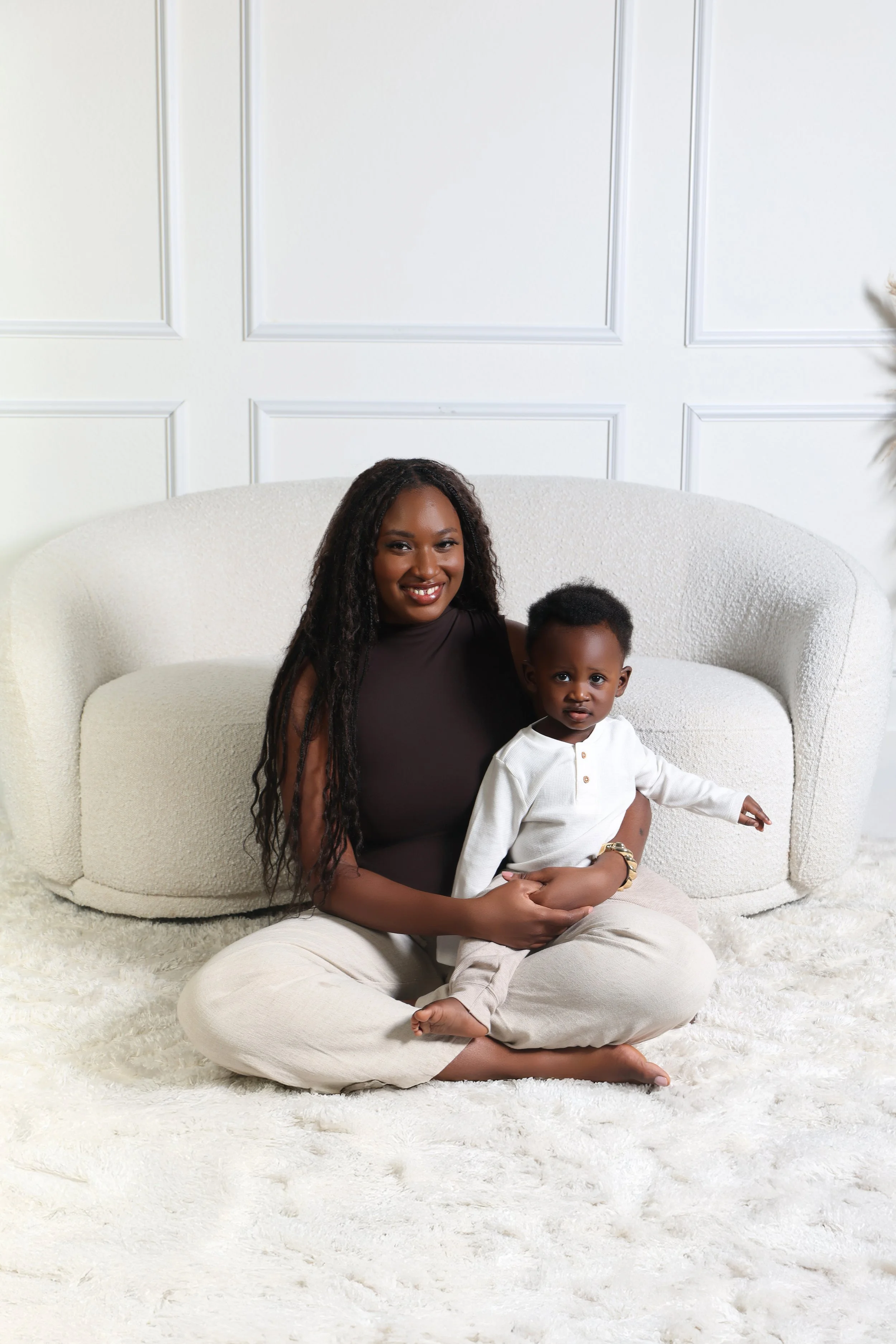A smiling woman with long curly hair sitting on a white textured rug in front of a cream-colored sofa, holding a young child with short curly hair wearing a white long-sleeve shirt and beige pants, inside a bright room with white paneled walls.