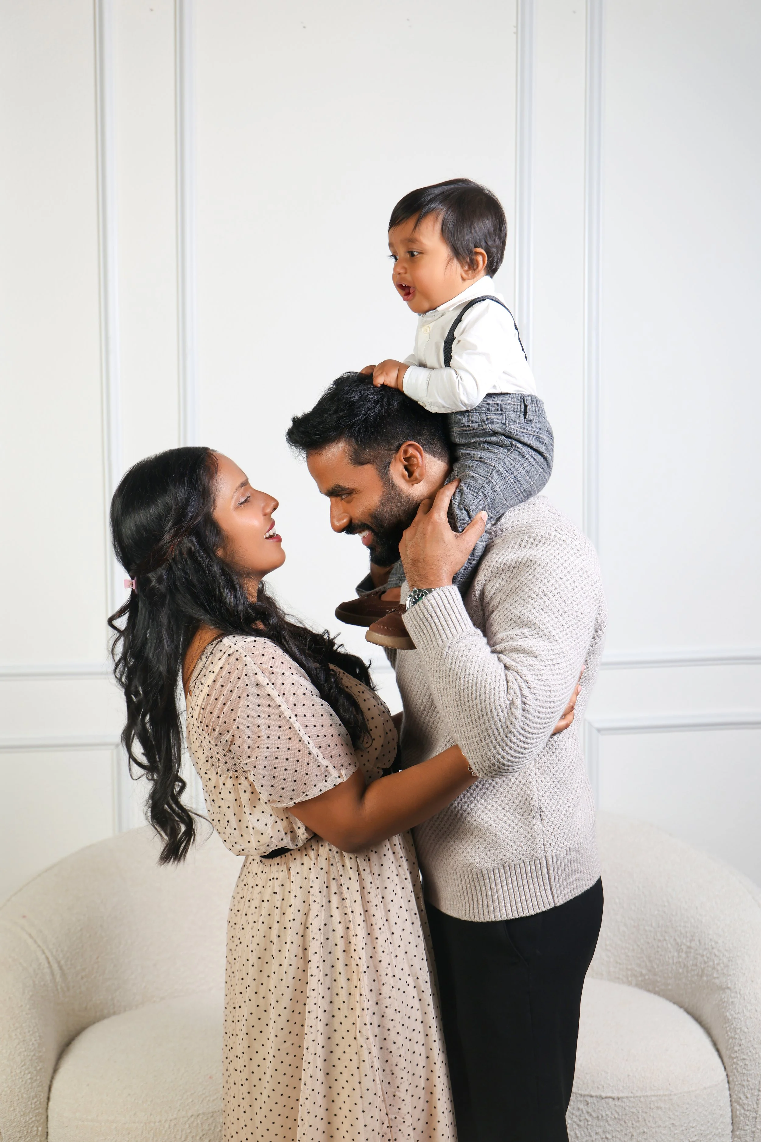 A happy family, a woman and two men, sharing a joyful moment inside a home. The woman and a man are smiling at each other while the man has a young child on his shoulders. The child is smiling and looking at the woman.