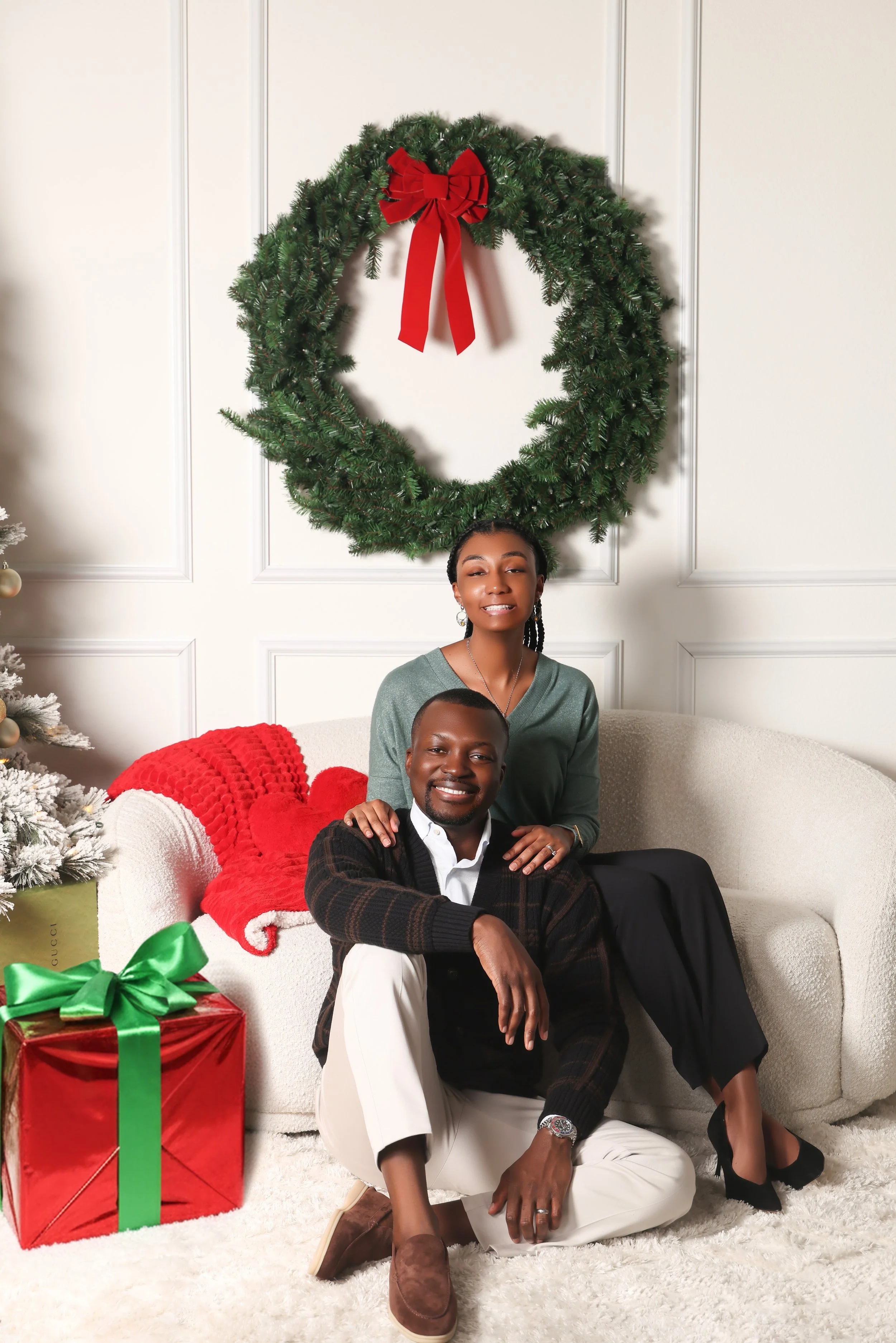 A couple sitting on a cream-colored couch in a festive Christmas setting, with a large green wreath with a red bow above them, a Christmas tree decorated with ornaments to the left, and a wrapped gift box with a green bow on the floor.