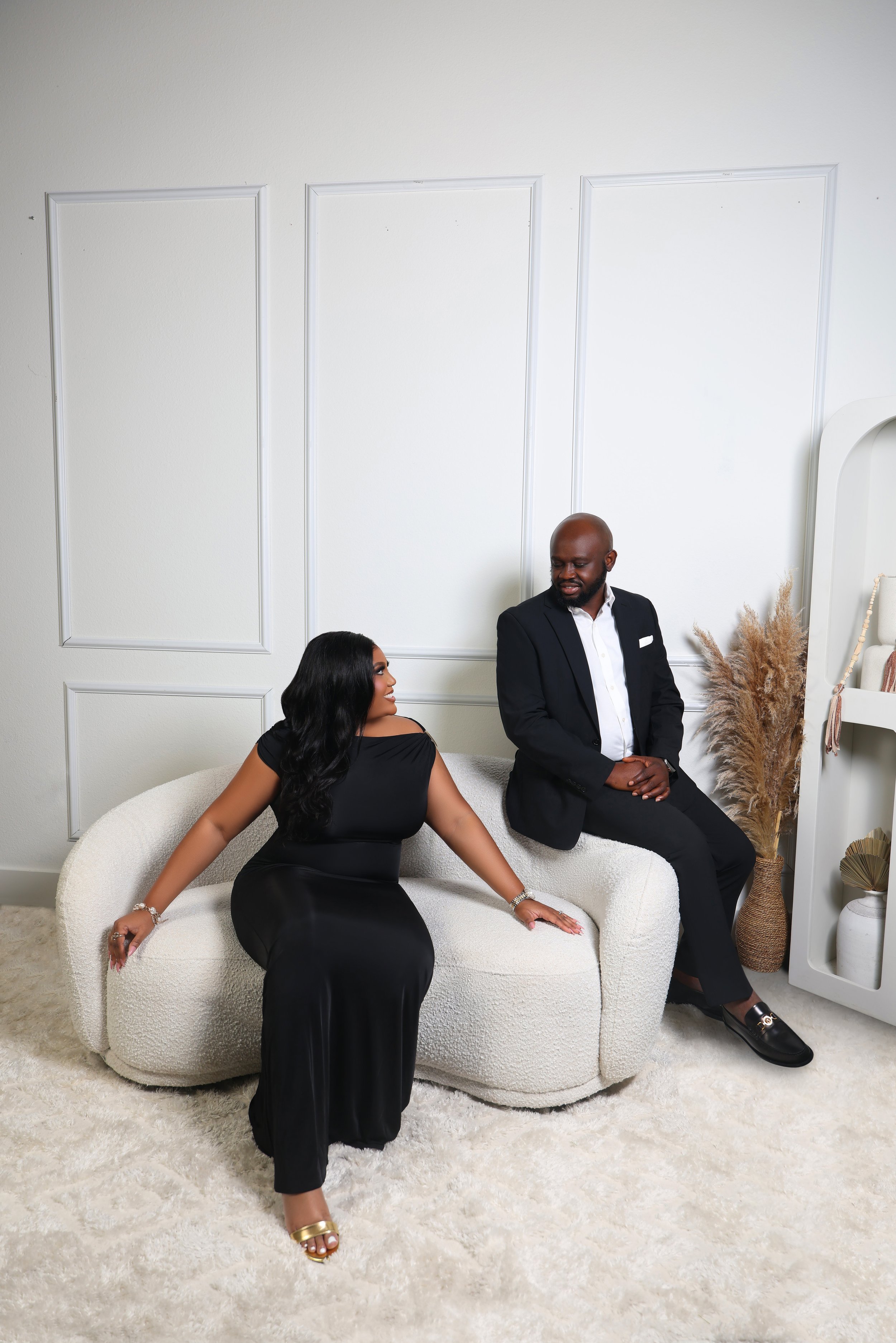 A woman and a man sitting on a white couch in a modern, minimalistic room with white walls and beige carpet. The woman is wearing a black dress and gold high heels, the man is in a black suit with a white shirt.