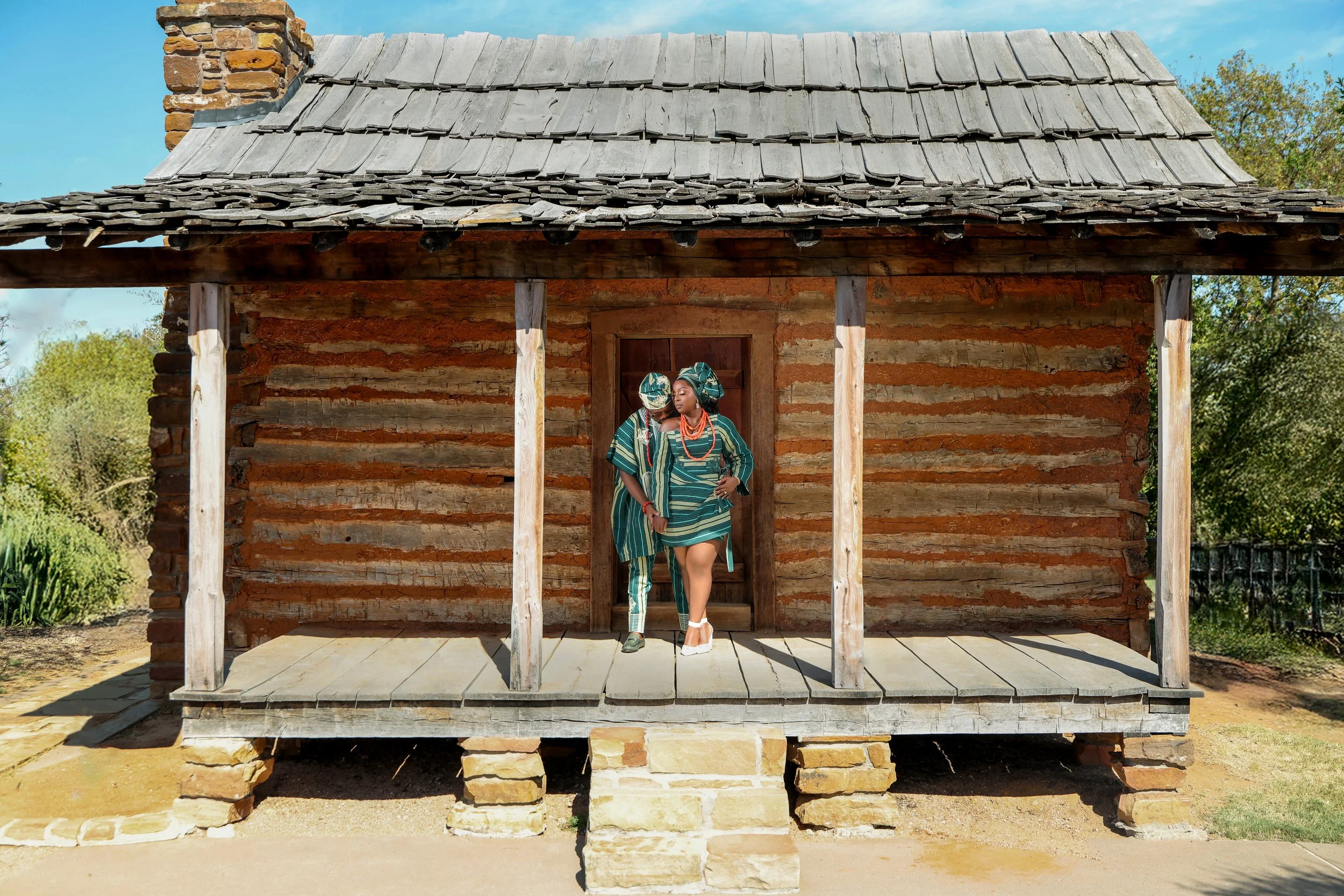Two people dressed in traditional African attire standing on a wooden porch in front of a small rustic log cabin with a shingled roof, surrounded by greenery under a blue sky.