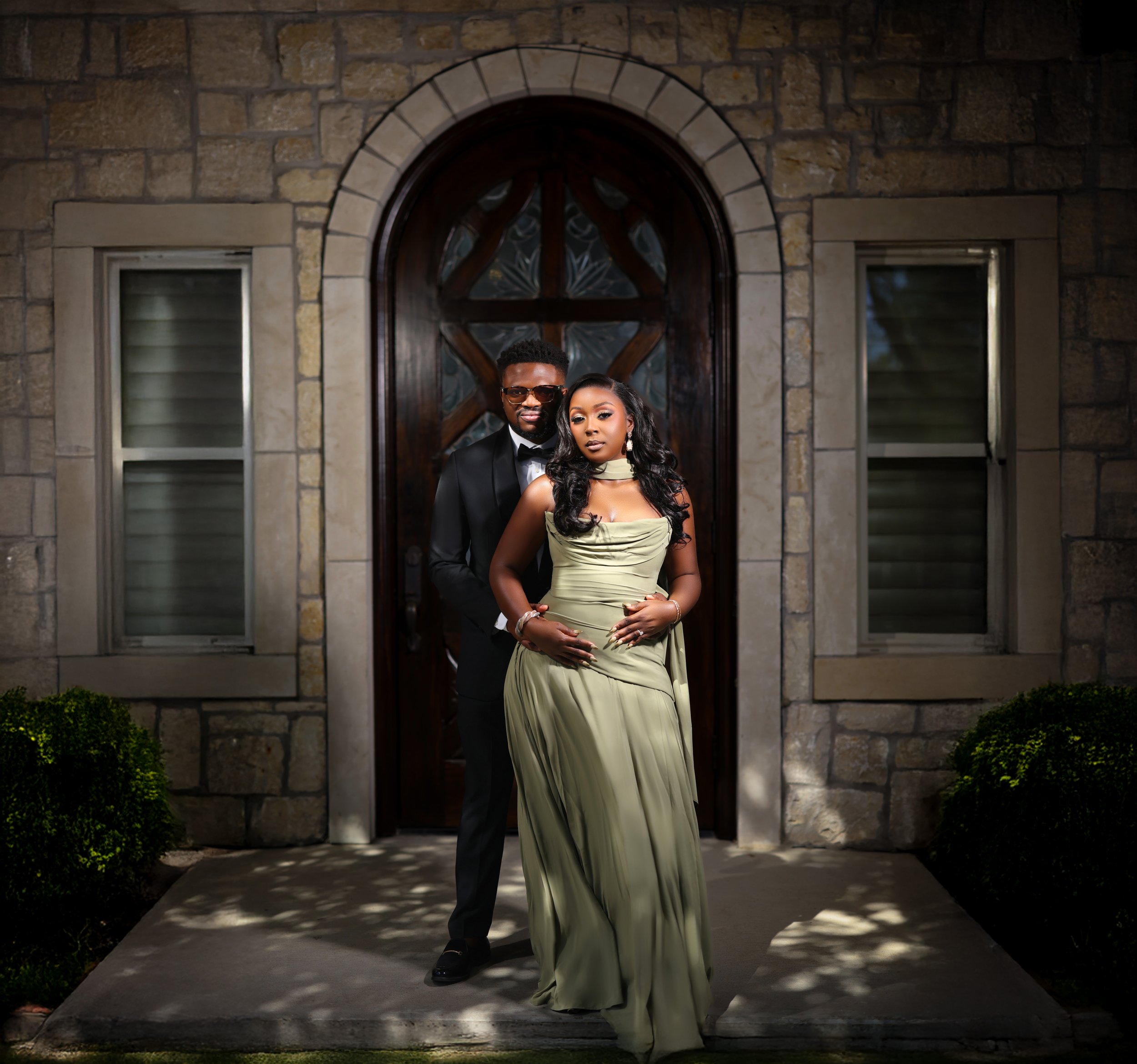 A stylish man and woman dressed in formal attire standing in front of a stone house with a wooden arched door and two windows.