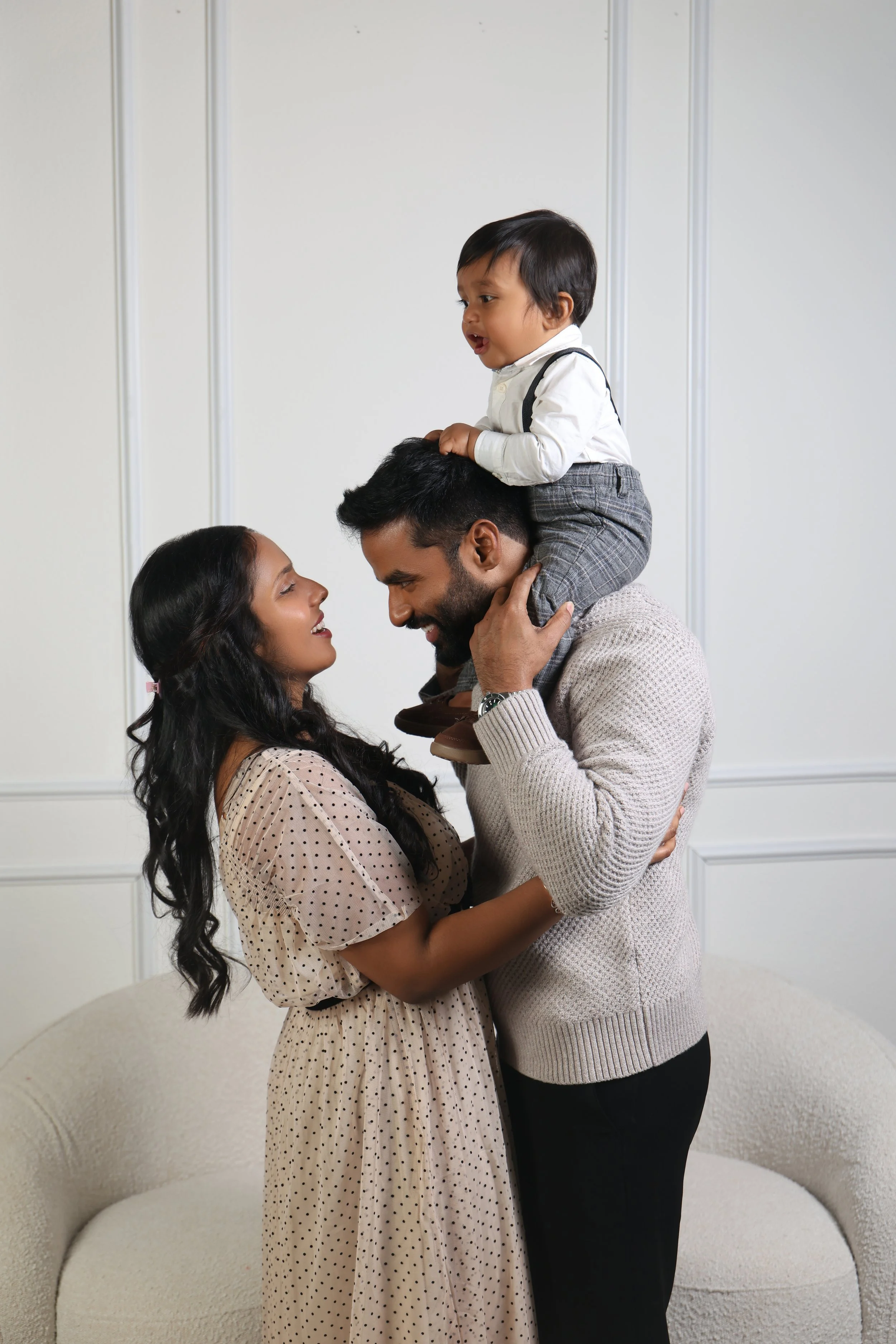 A family of three, an Asian woman, an Indian man, and a young boy, enjoying a playful moment together indoors. The boy is sitting on the man's shoulders, and the woman is holding the man's waist, all smiling and looking at each other.