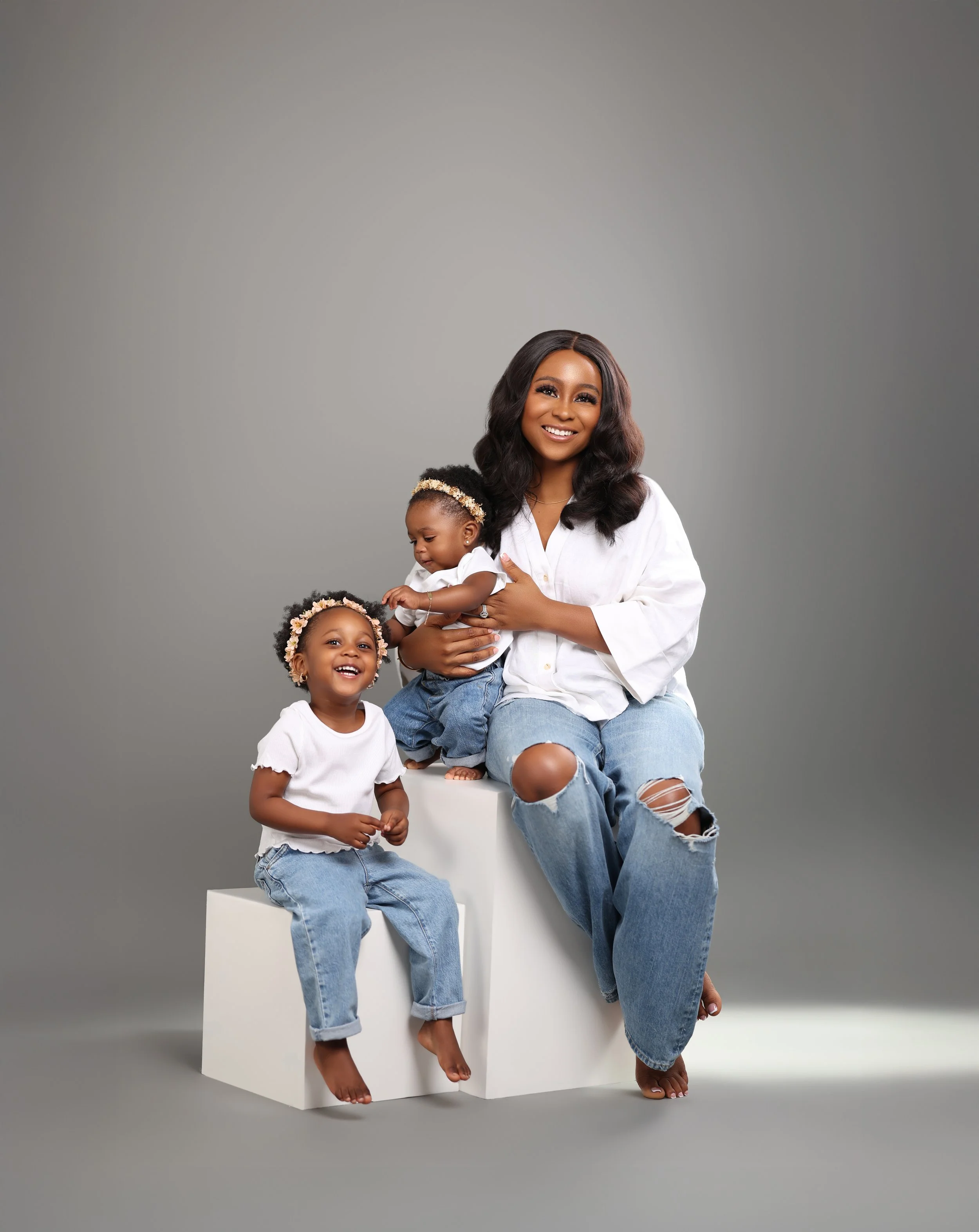 A smiling woman sitting on white cubes with two young girls, all wearing casual white shirts and jeans, in a studio with a gray background.