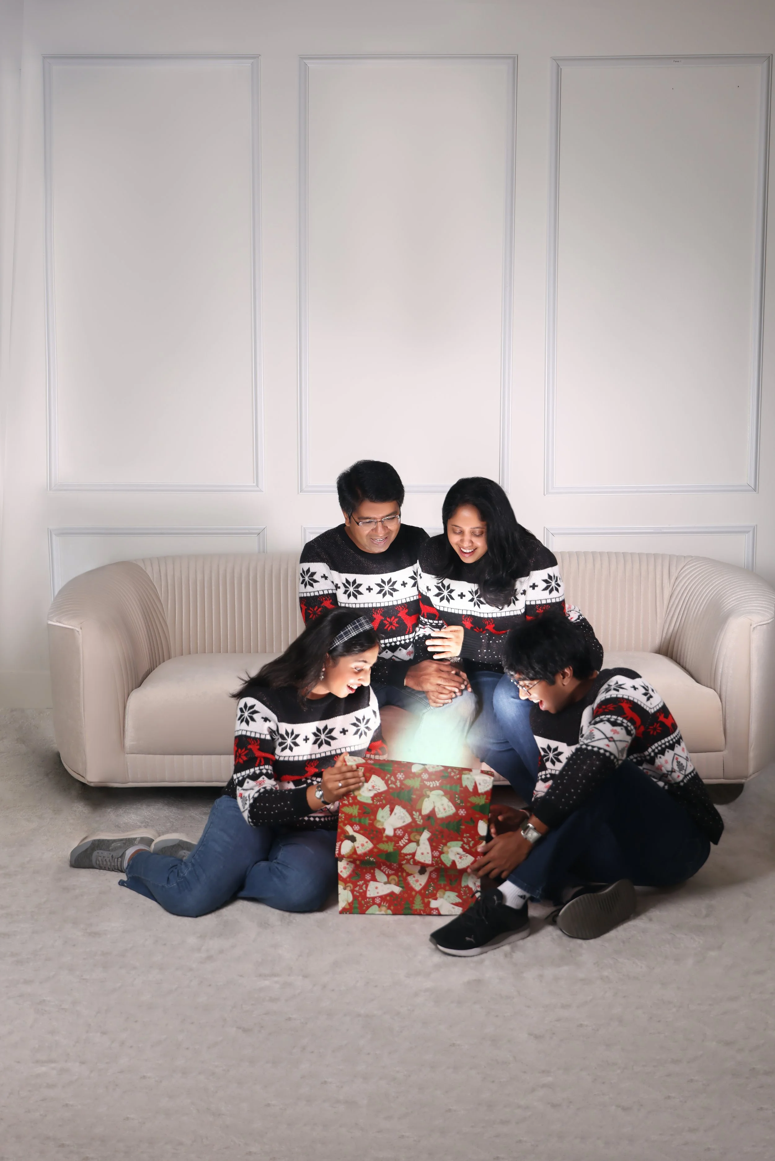 A family of four in matching holiday sweaters opening Christmas presents inside a living room.