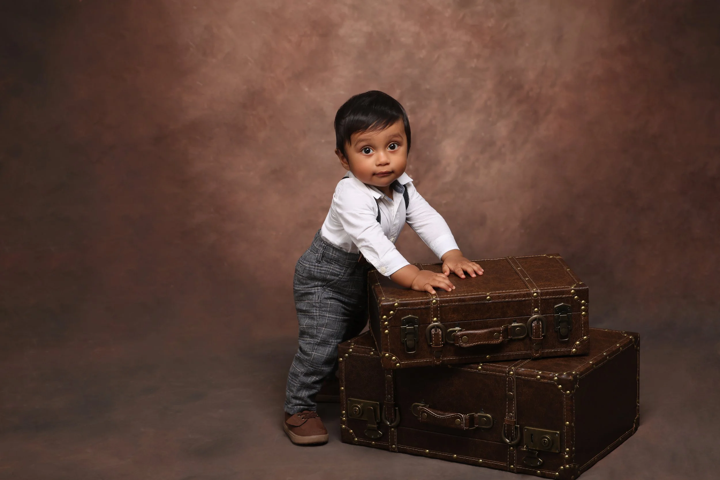 Young boy in white shirt and plaid pants standing behind stacked vintage brown suitcases.