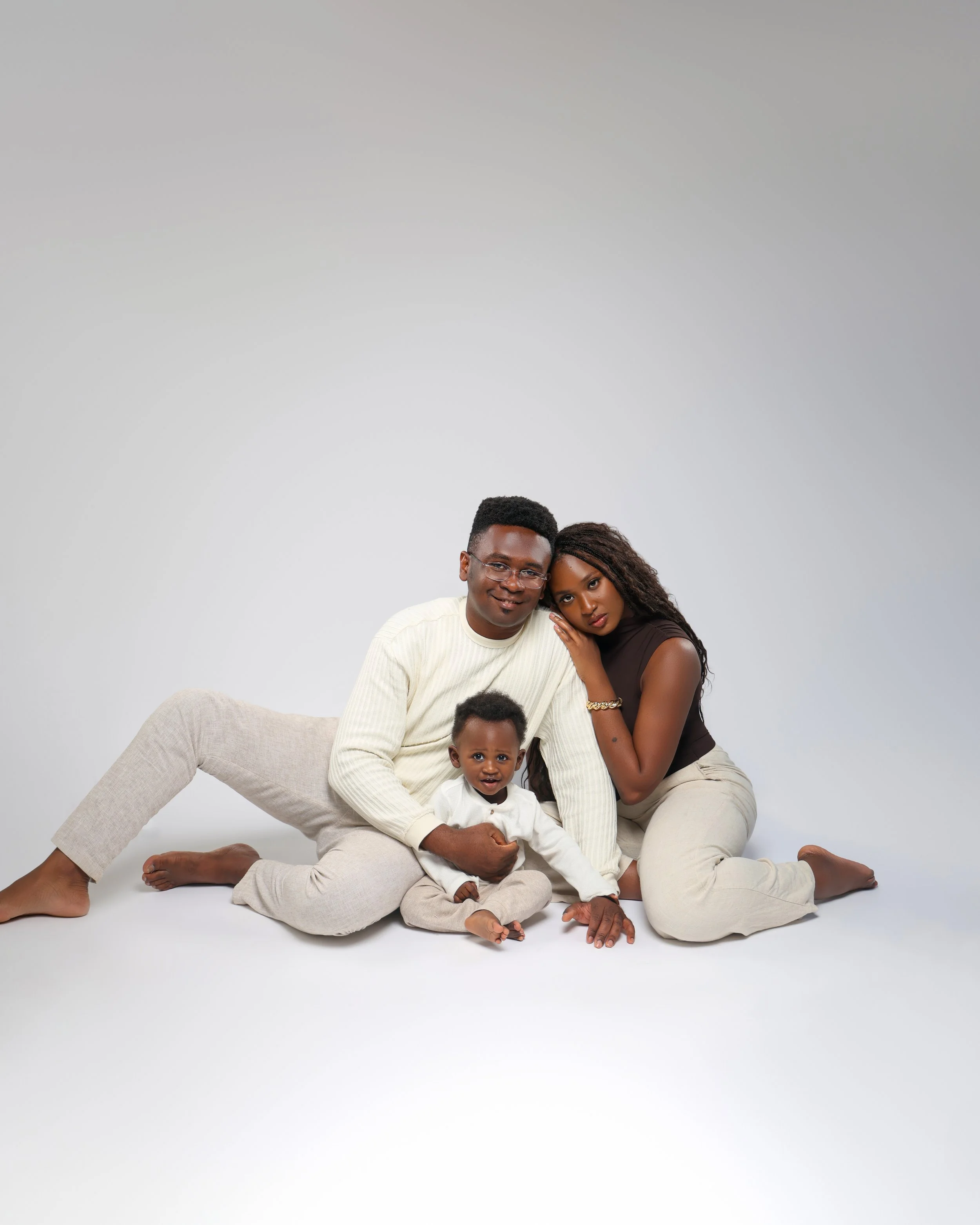 Family of three posing on the floor against a white background, with the father and mother sitting close and the child in front, holding hands with the father.