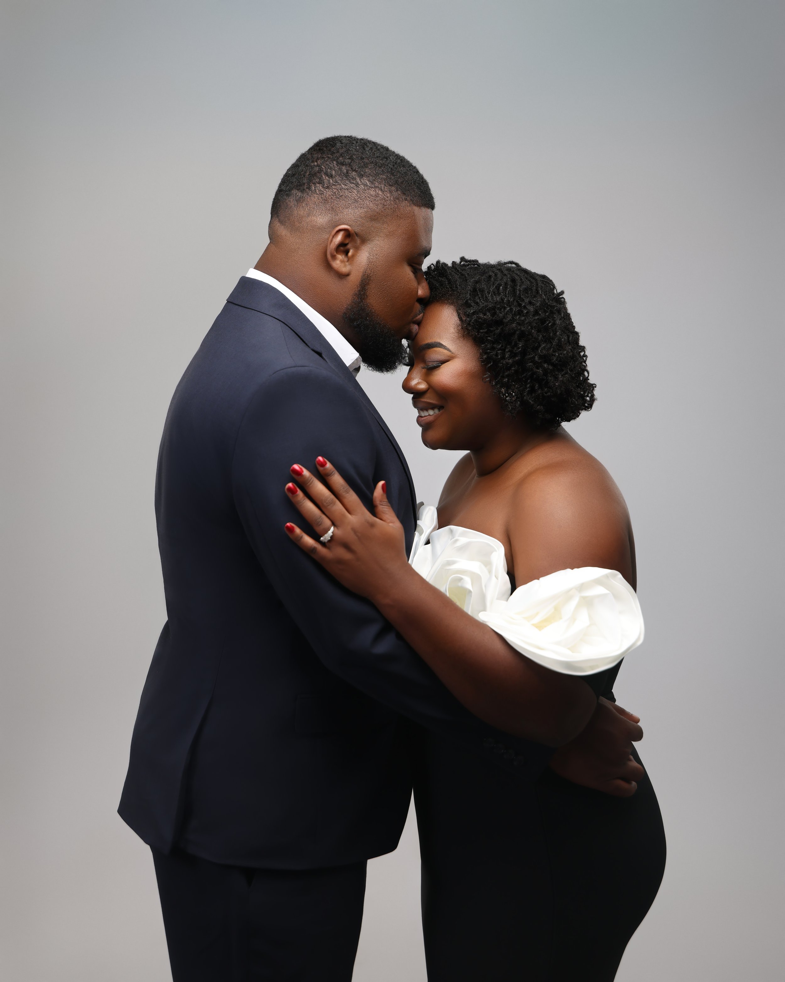 A couple in wedding attire, embracing against a plain gray background. The man is in a dark suit, and the woman is in an off-the-shoulder white dress with puffed sleeves, smiling with eyes closed.