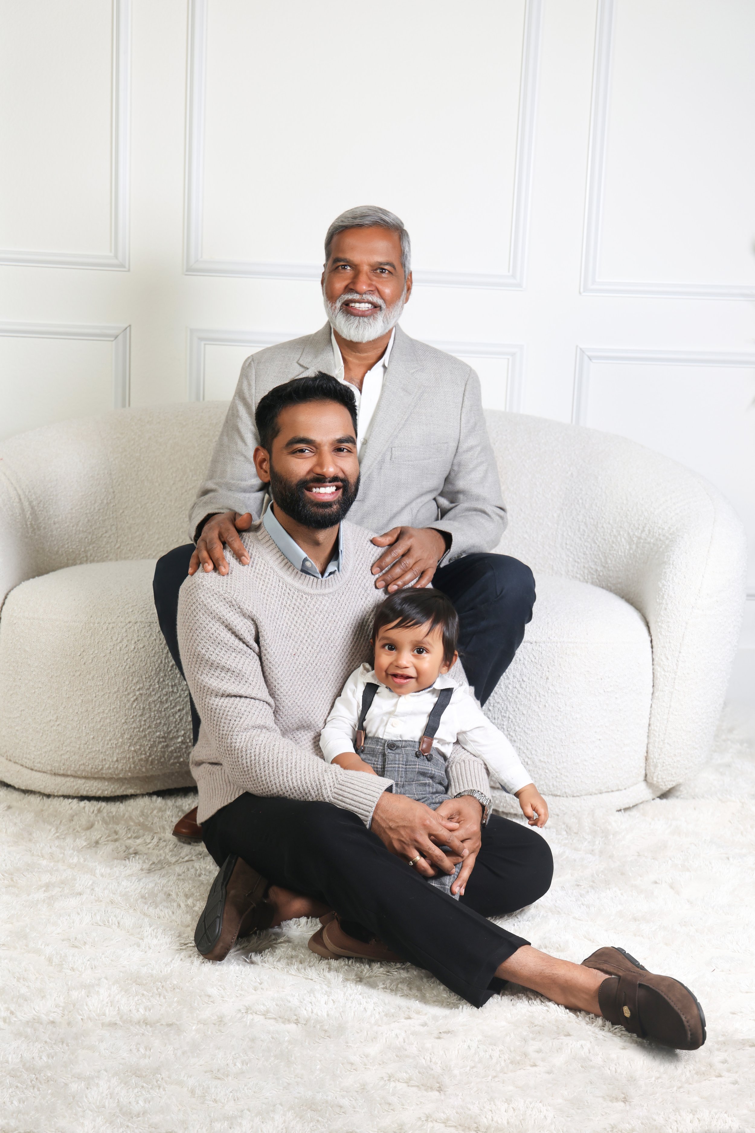An older man, a younger man, and a young child sitting on a white carpet in a living room with white paneled walls, smiling at the camera.