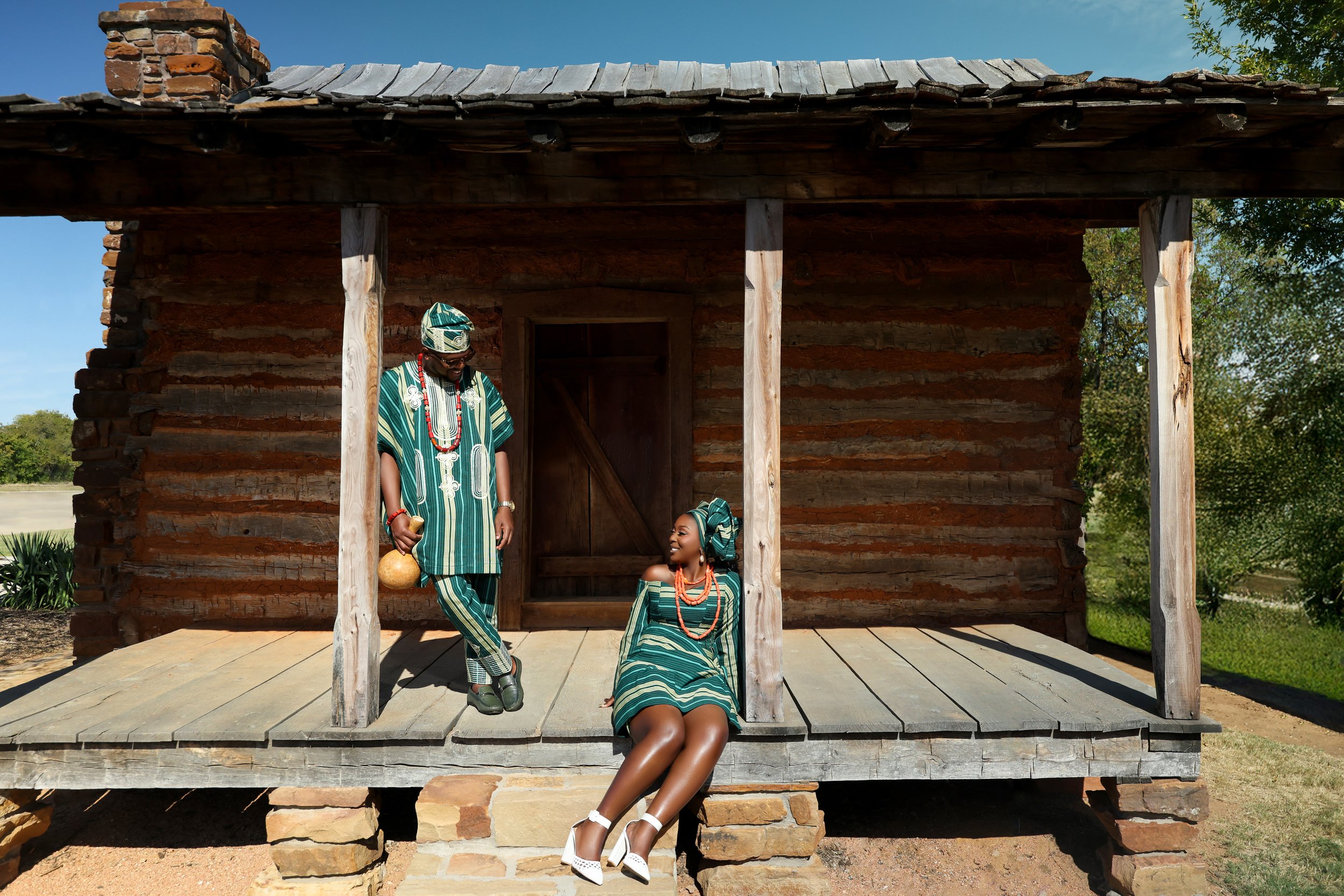 A man and a woman dressed in traditional African attire on a rustic wooden porch of a small stone and wood cabin, with the woman sitting and the man standing nearby, both smiling at each other.