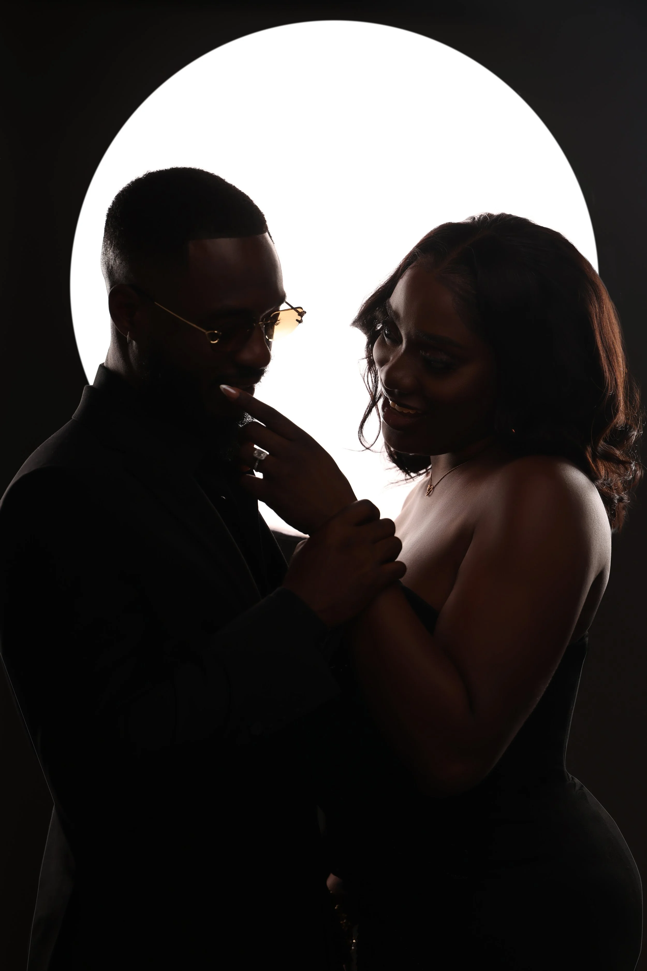 Silhouetted man and woman close together, illuminated from behind by a large circular light, creating a romantic mood.