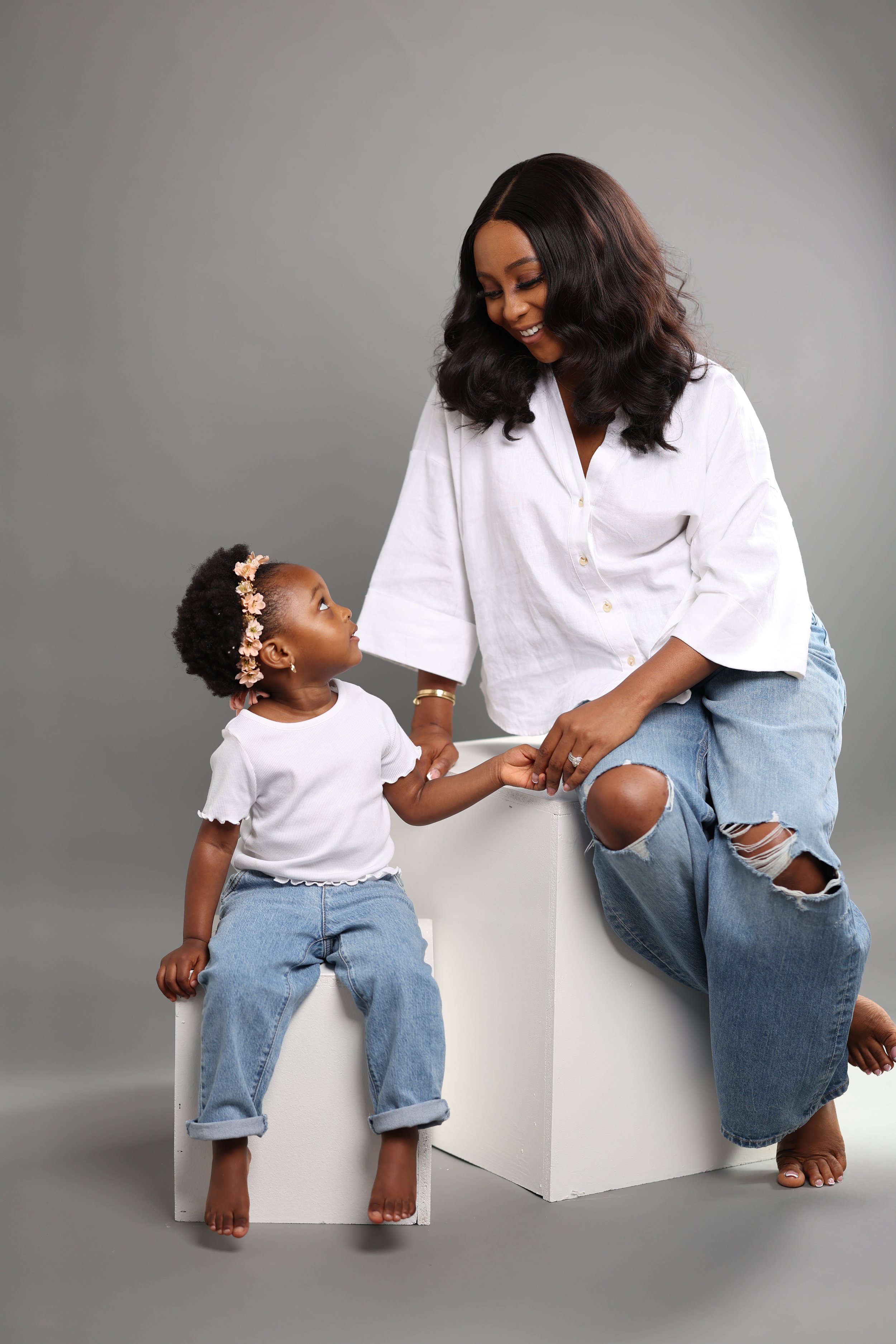 A woman sitting on a white block, smiling at a young girl who is sitting on a smaller white block, holding her hand and looking up at her. Both are dressed casually in white tops and ripped jeans, with the girl wearing a floral headband. The backgrou