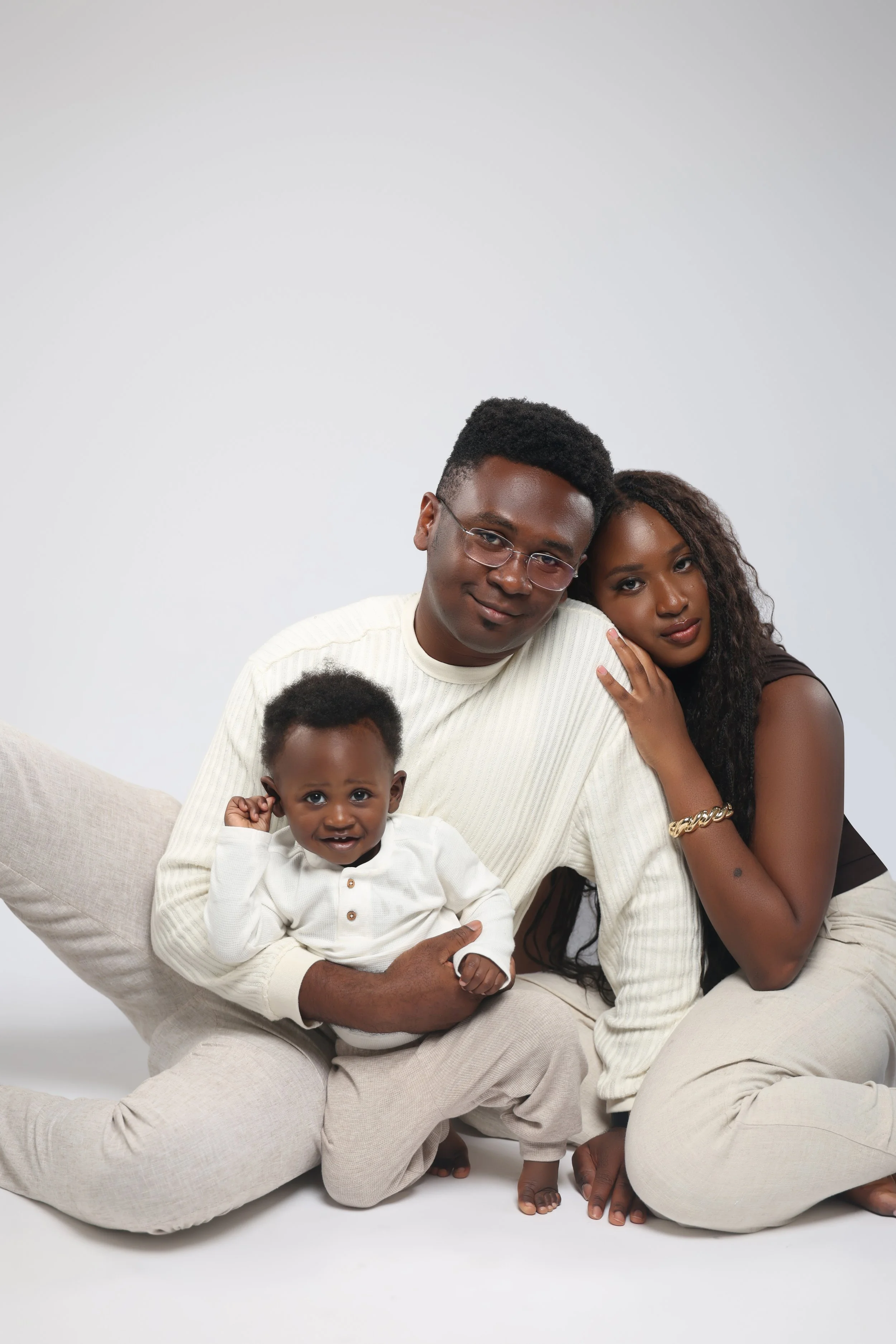 A family of three sitting on the floor against a plain gray background. An adult man with glasses, a woman, and a young child in white clothing.