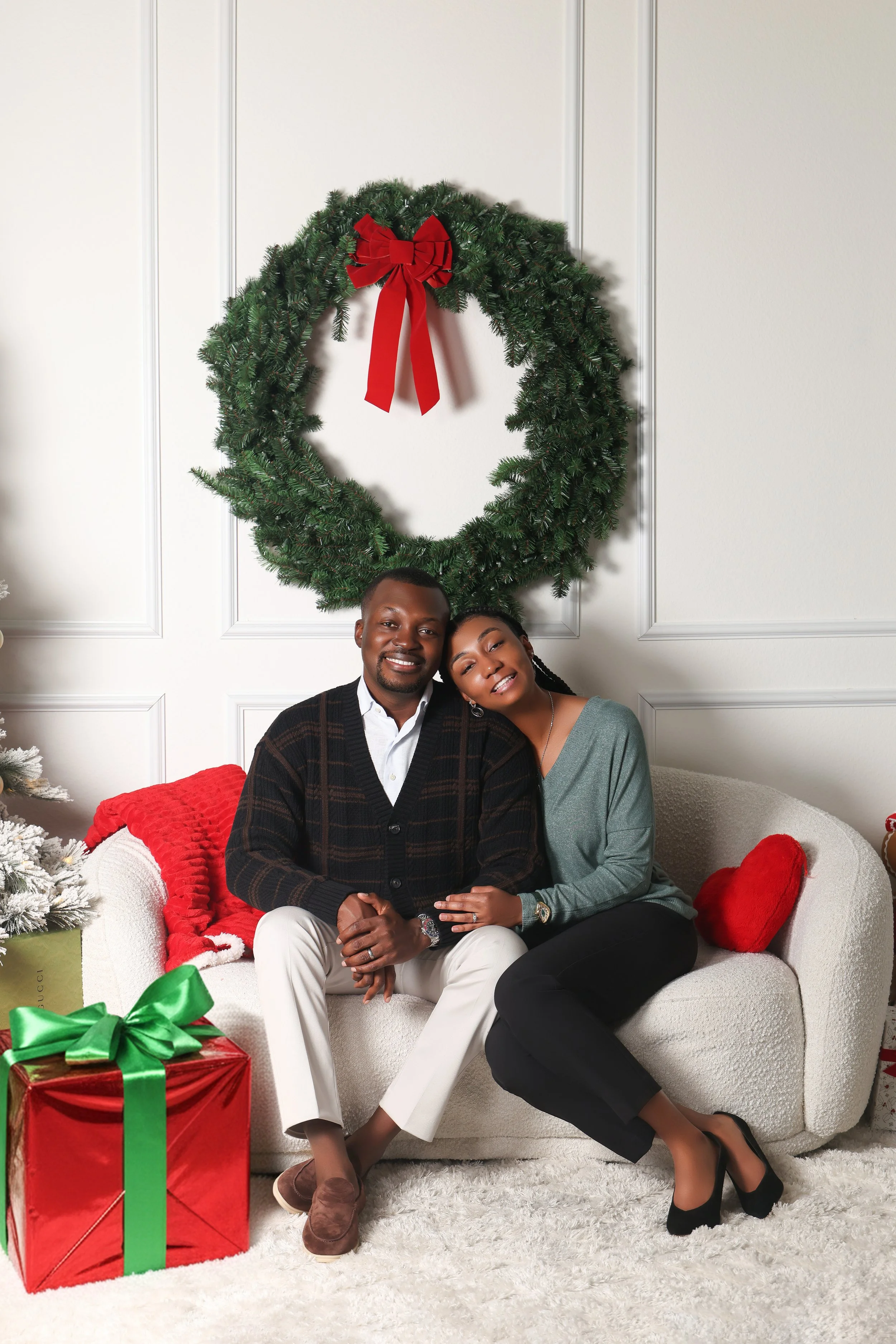 A smiling couple sitting on a white couch in a decorated room with a Christmas wreath, wrapped gift, and Christmas tree.