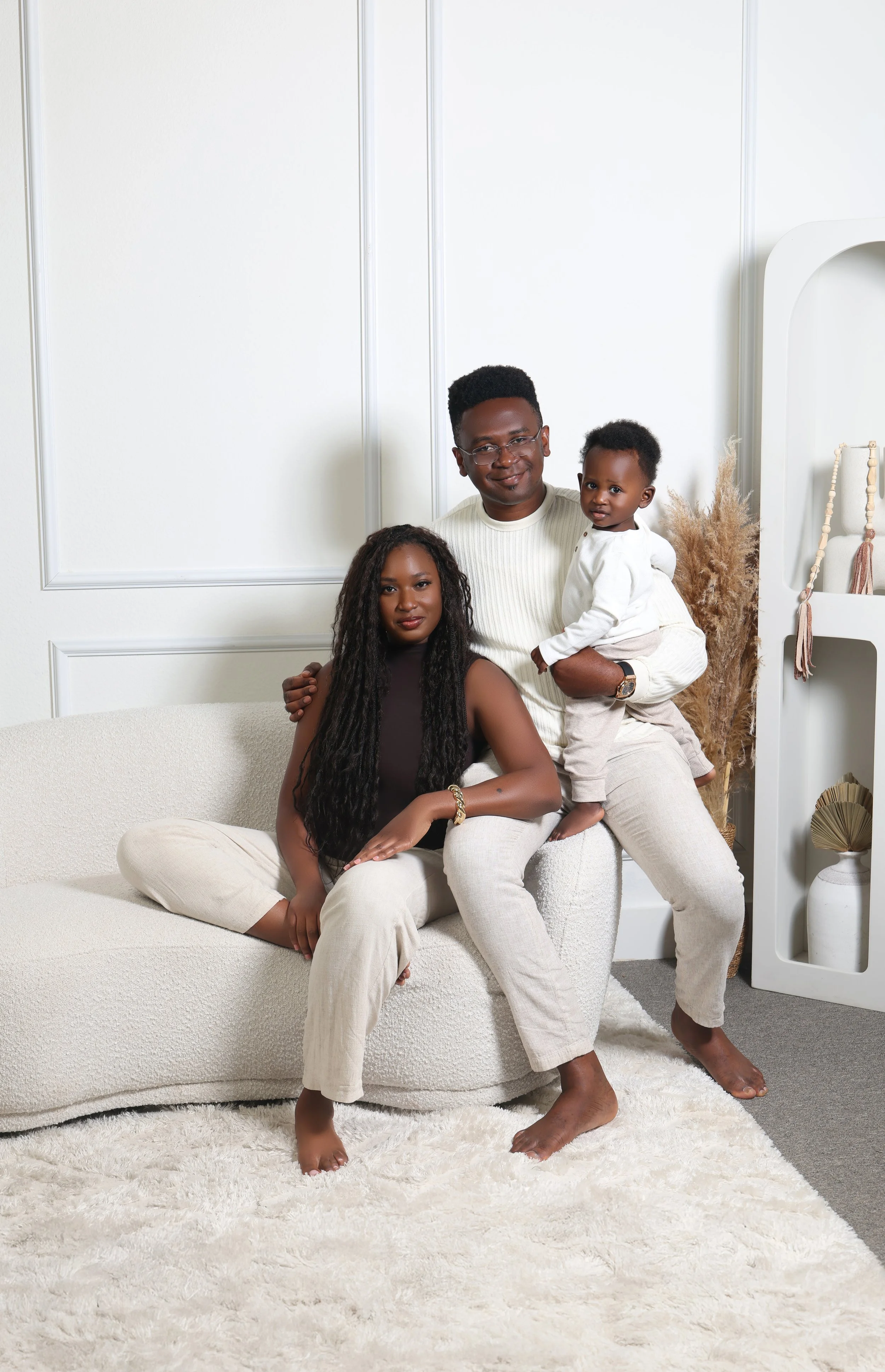 A family of three with dark skin in a bright, modern living room. The woman sits on a white sofa with long curly hair and wears a sleeveless top. The man, with short hair and glasses, stands behind her holding a young child. The child, in light-color