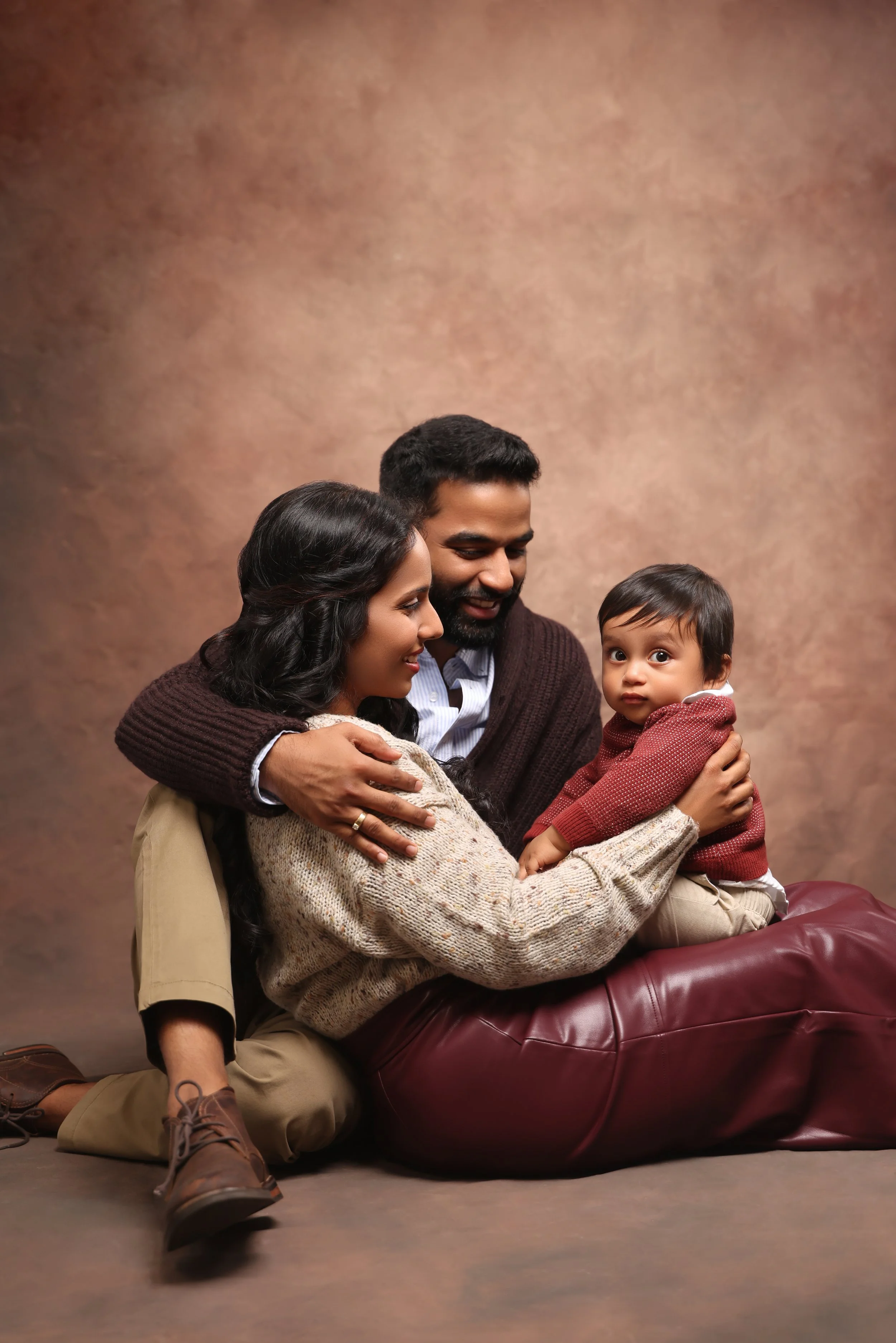 A happy family of three with a young child, sitting on the floor against a neutral background. The parents are embracing the child and smiling, the mother is sitting on the floor, the father is kneeling behind her, and the child is sitting on a maroo