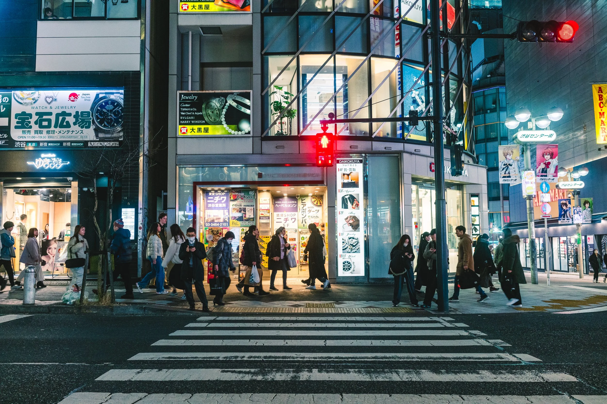 A city street scene at night with pedestrians crossing a crosswalk in front of a modern building with various illuminated signs and advertisements in Japanese. The traffic light is red.